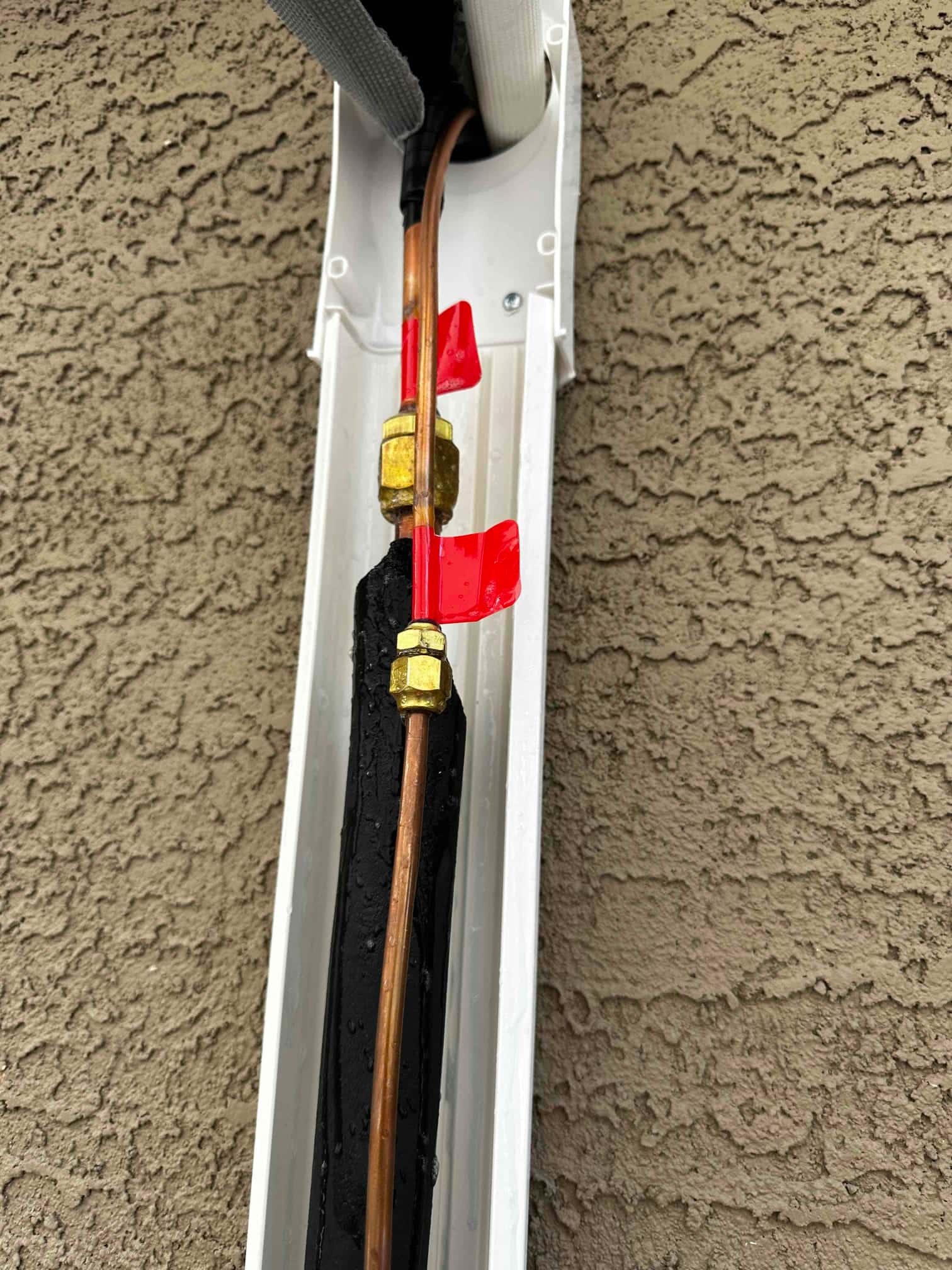 Refrigerant line set with flare unions routed in a protective line cover on exterior wall, insulation and drain present.
