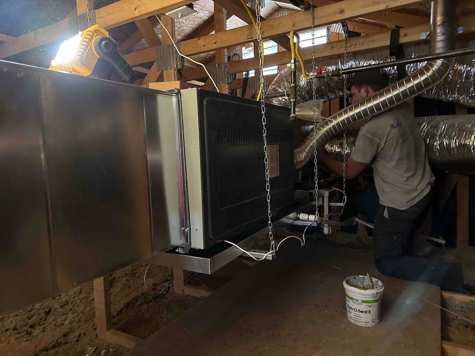Technician works in attic installing an air handler with metal plenum, insulated flex ducts, hanging chains, and condensate drain piping.