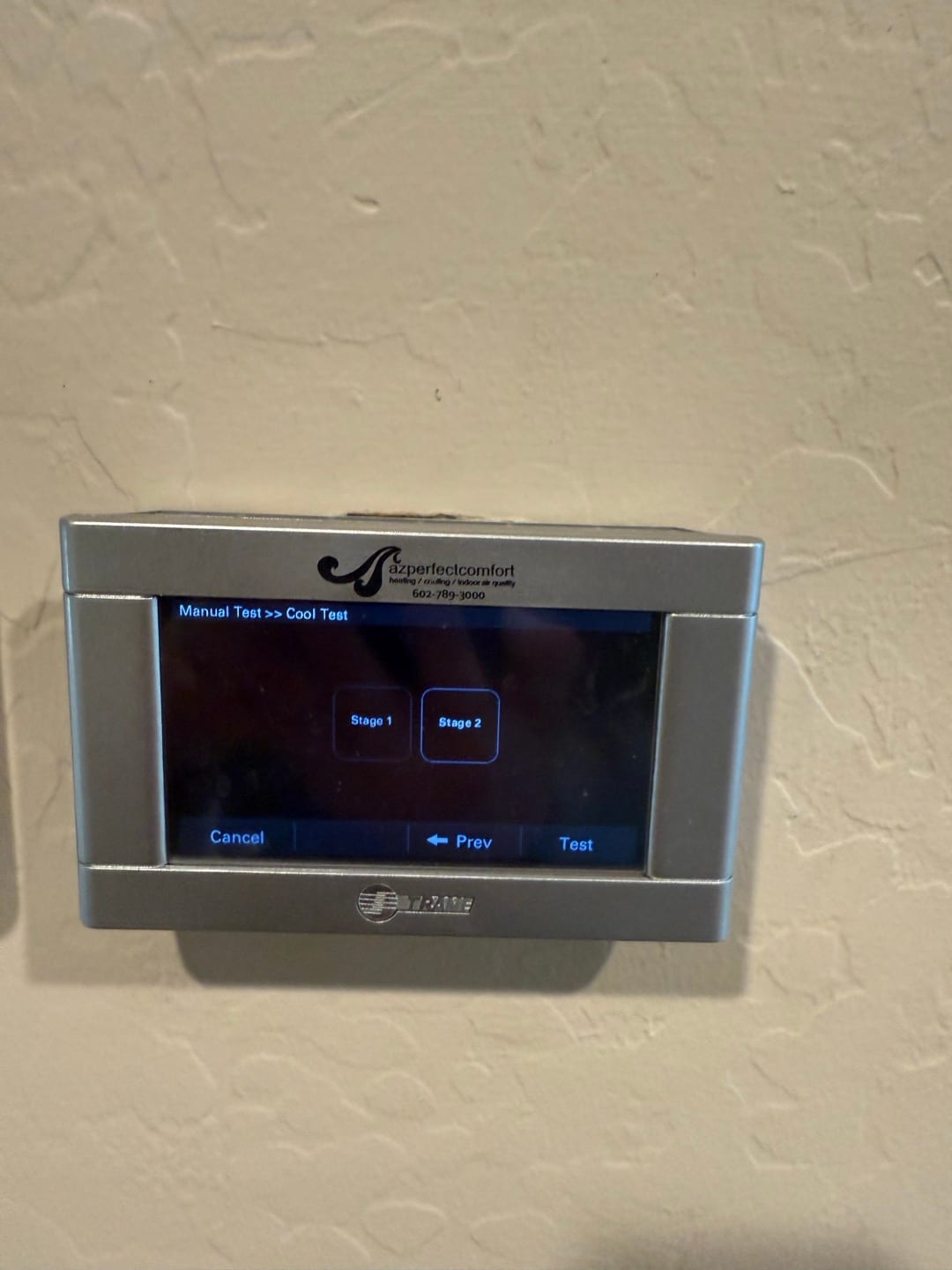 Touchscreen thermostat on wall displaying manual cooling test menu with options for Stage 1 and Stage 2.