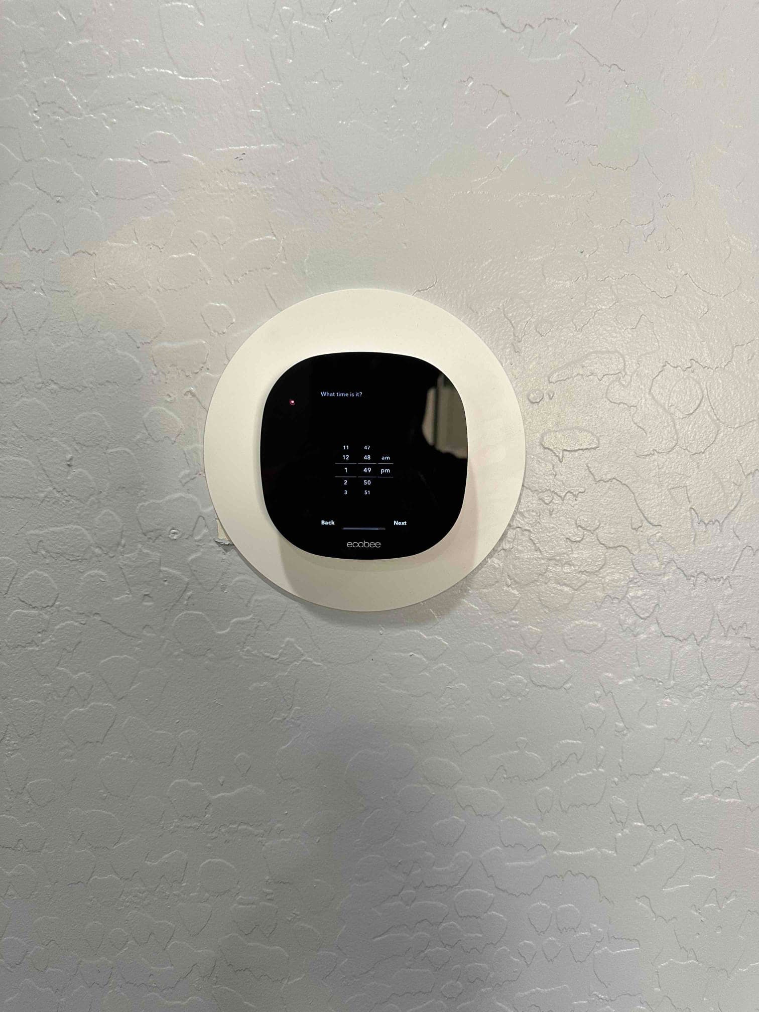 Wall-mounted smart thermostat installed with trim plate, powered on and prompting for time setup during commissioning.
