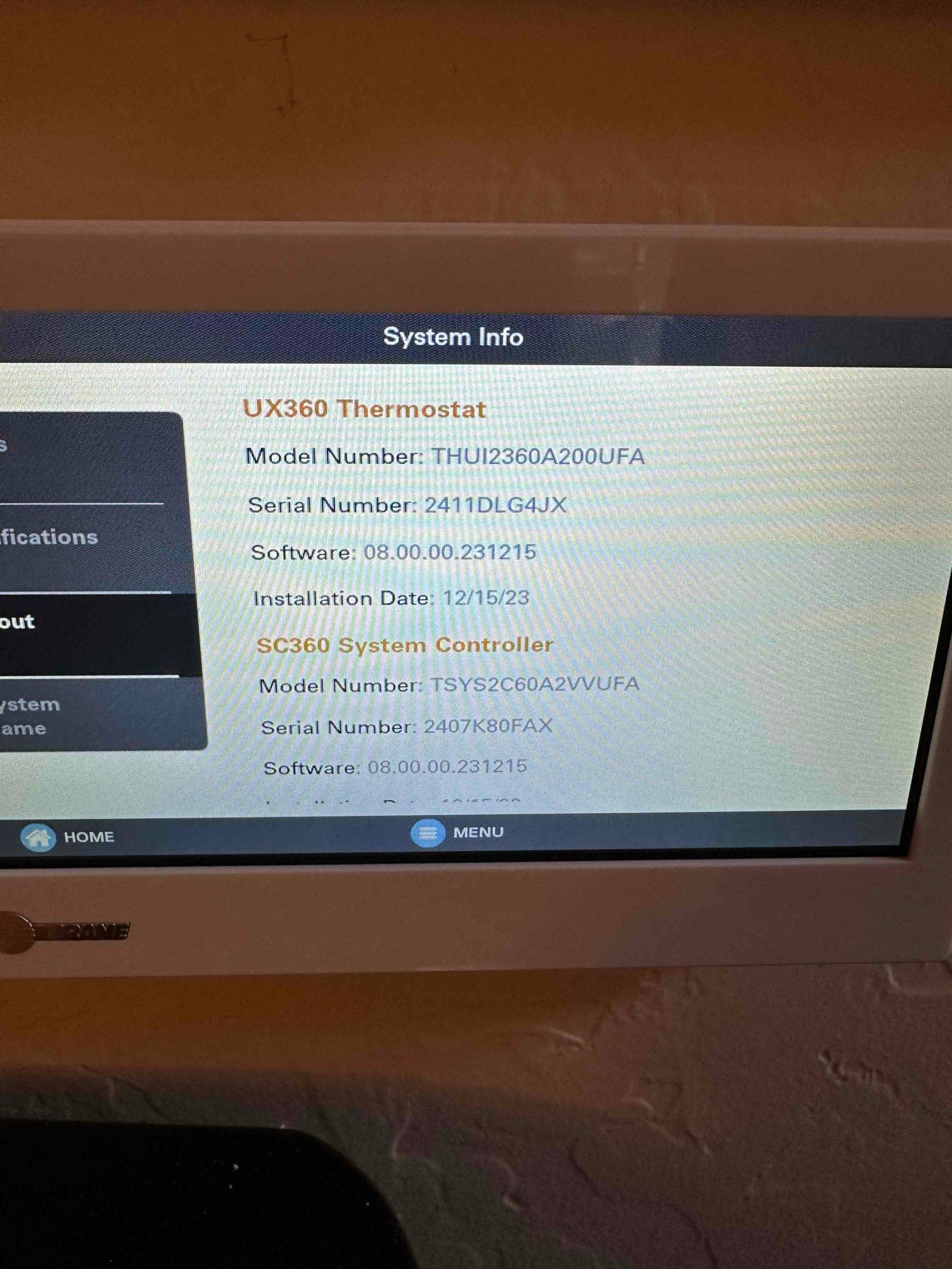 Wall-mounted touchscreen thermostat displays system information screen with controller details and installation date visible.