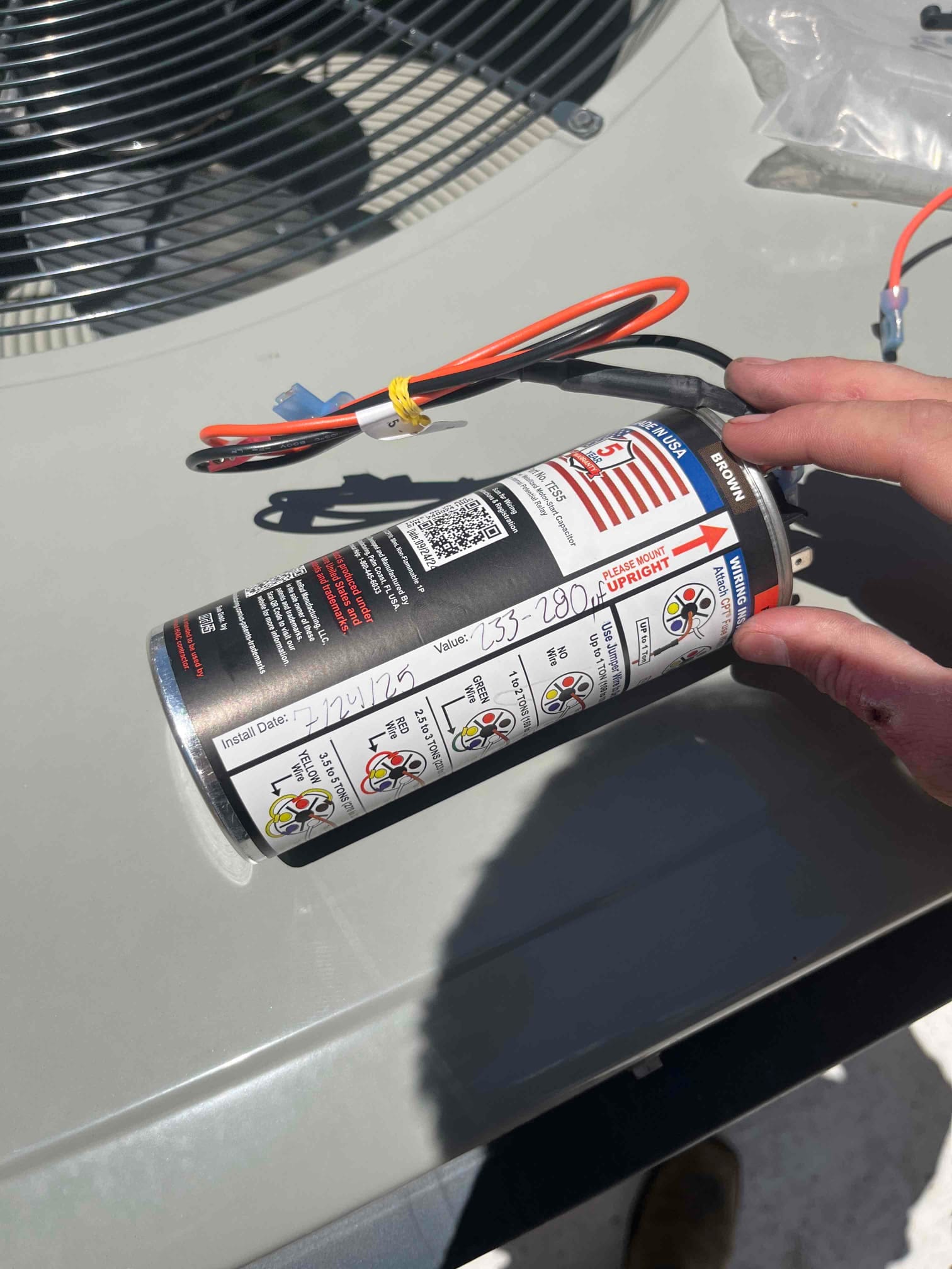 Hard-start capacitor kit held near the outdoor condenser, showing wiring leads and printed connection diagram on the label.