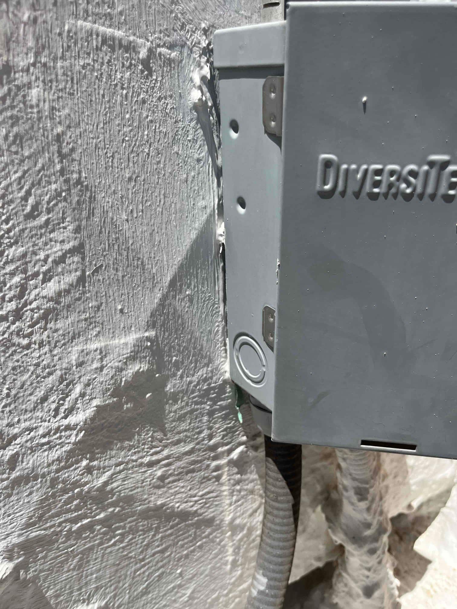 Outdoor disconnect box with flexible conduit entering the enclosure next to a textured exterior wall.