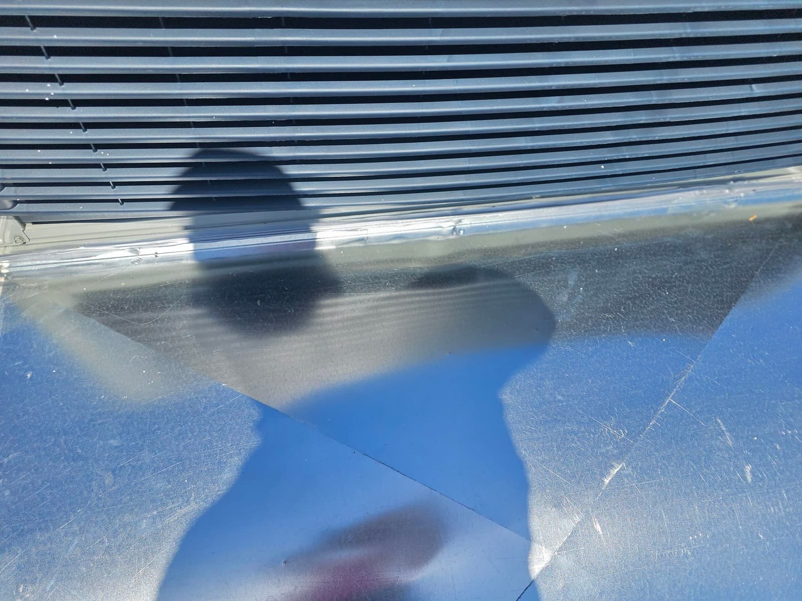 Sheet metal curb or flashing beneath outdoor HVAC unit grille shows clean seams and reflective surface in sunlight.