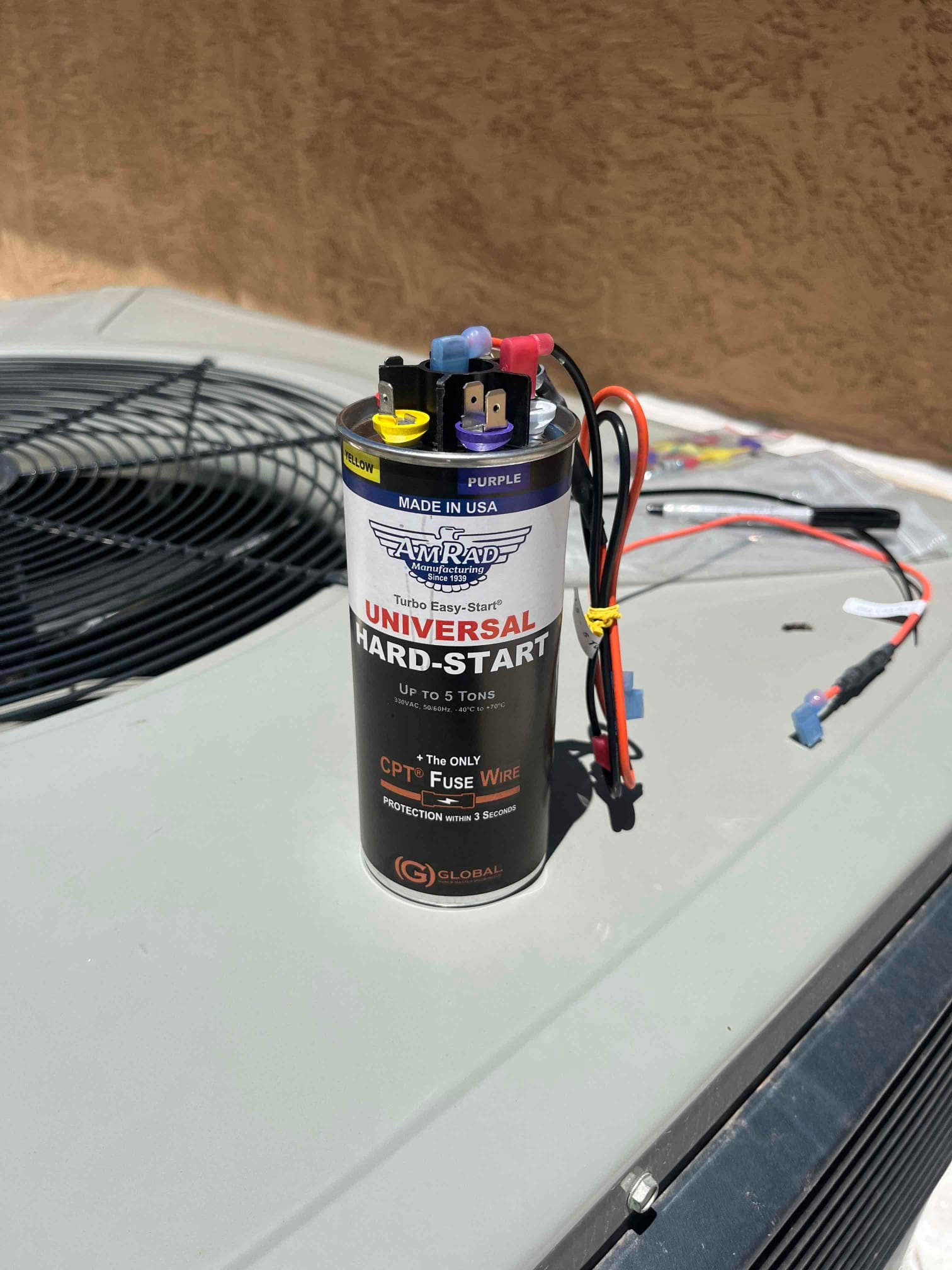 Universal hard-start kit with wires and terminals rests on top of an outdoor condenser unit in sunlight.