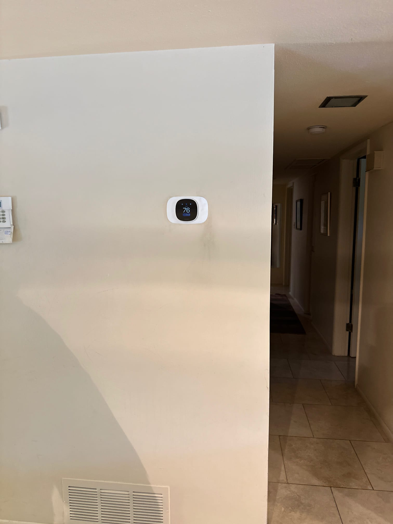 Wall-mounted smart thermostat installed and powered on near a return grille in the hallway area.