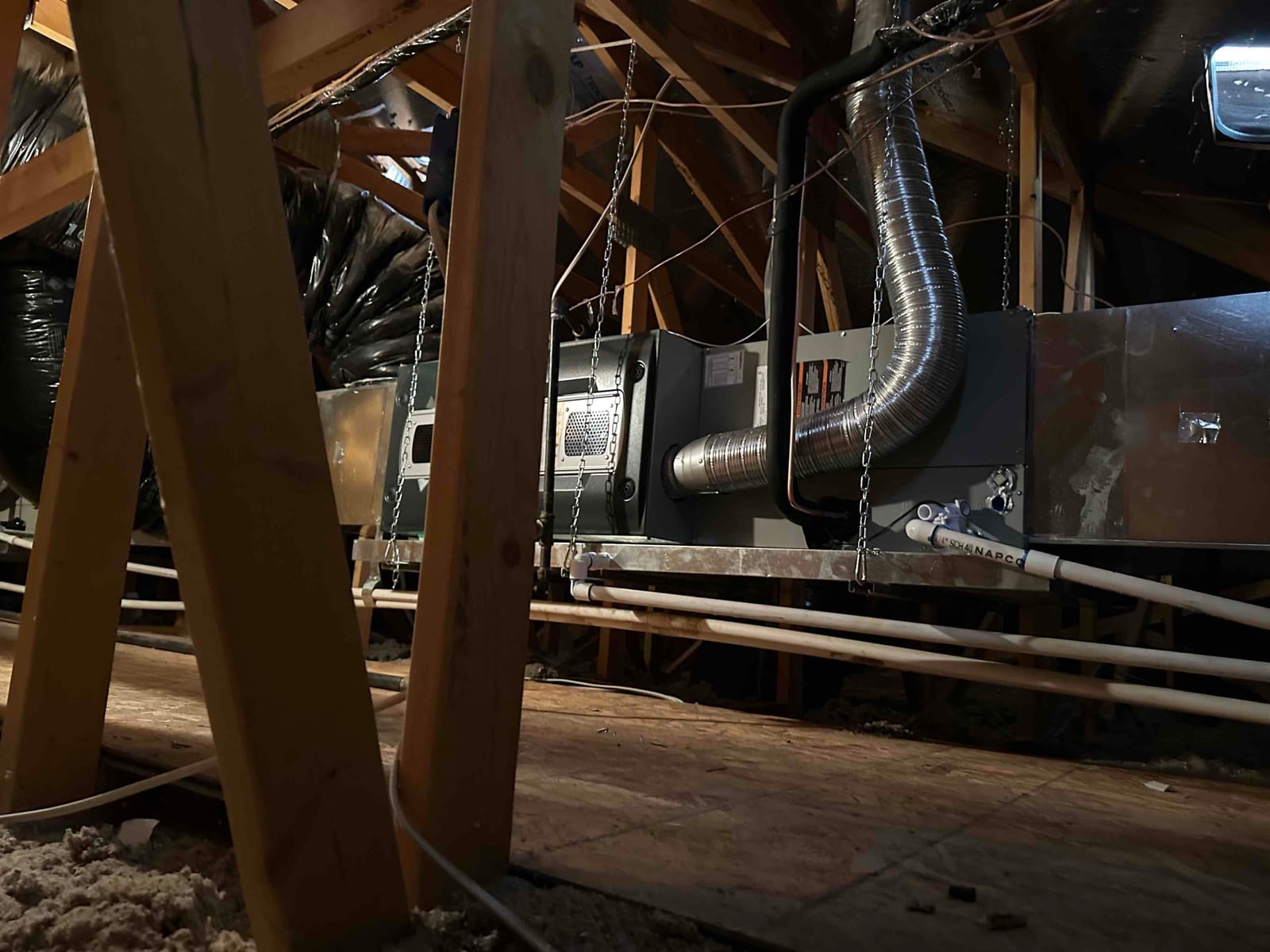 Air handler and ductwork suspended in attic with insulated refrigerant line, metal vent, drain piping, and support chains installed.
