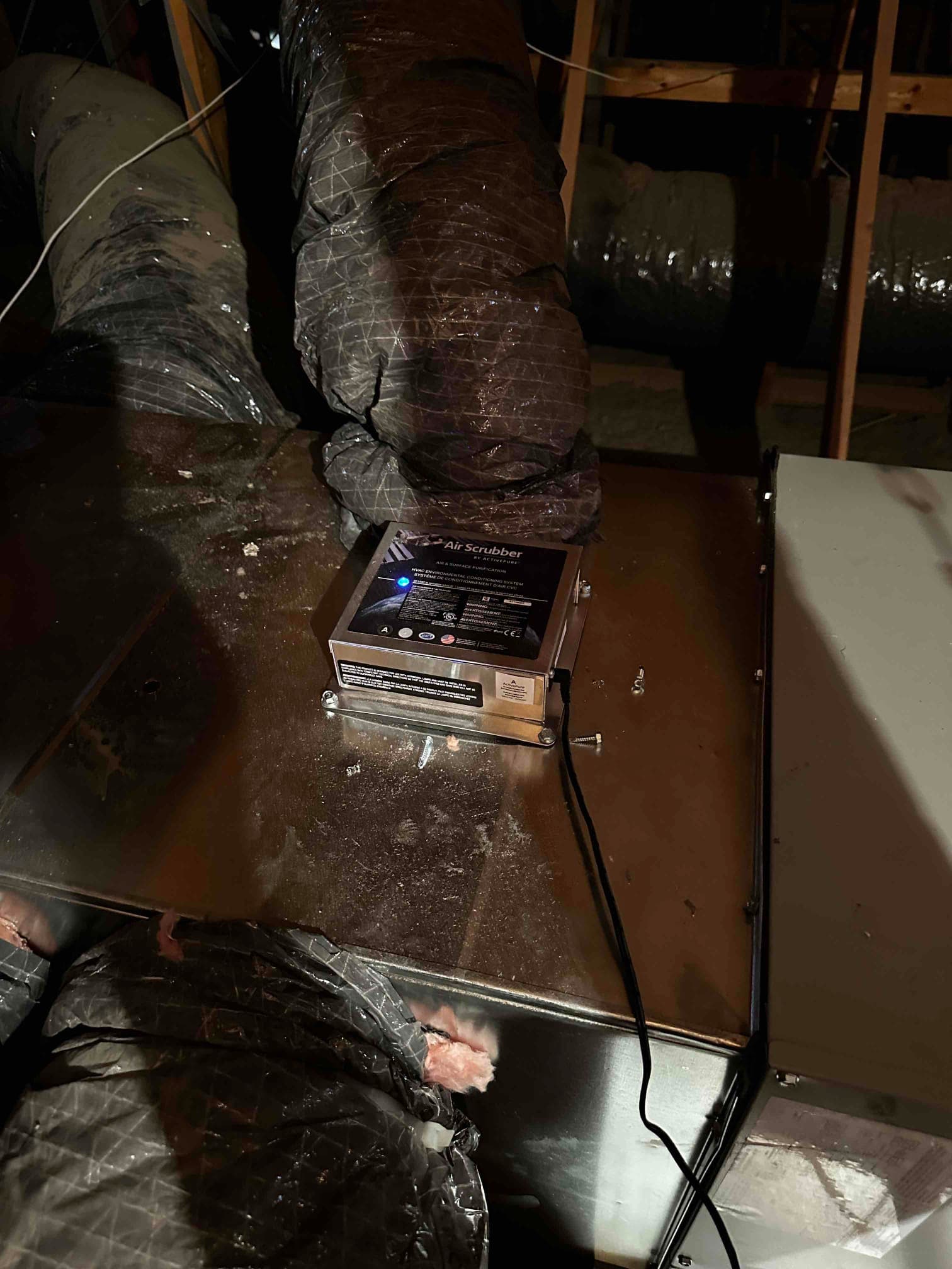 Air purification device mounted on the supply plenum with flexible duct connections visible in the attic space.