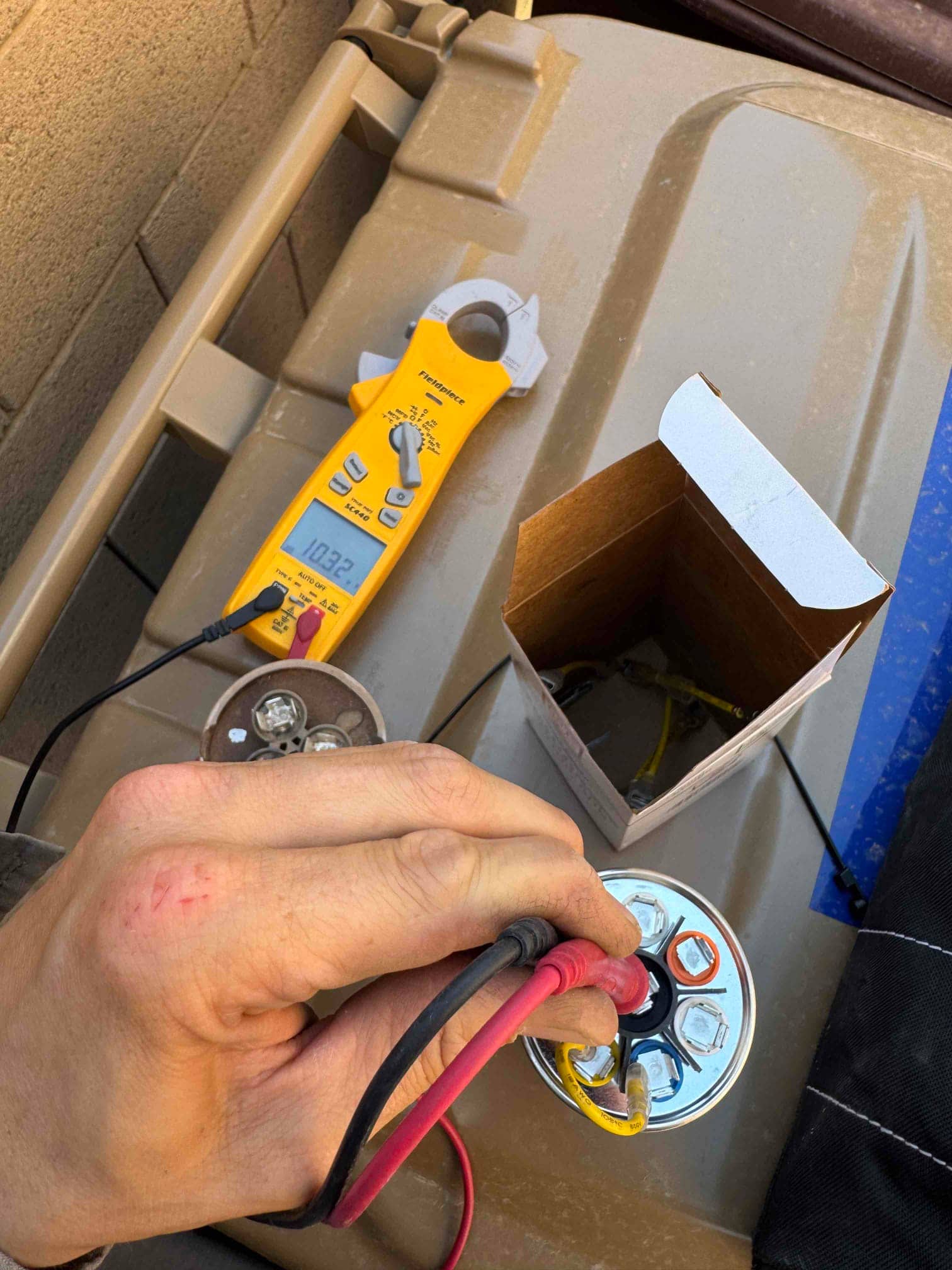 Multimeter measures 10.32 μF on the run capacitor while leads contact its terminals during air conditioner repair.