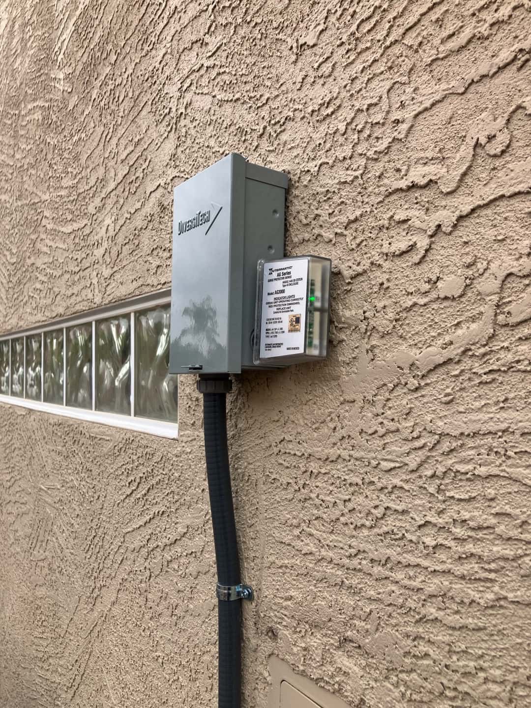 Outdoor HVAC disconnect with flexible conduit and surge protection installed cleanly on exterior wall beside glass block window.