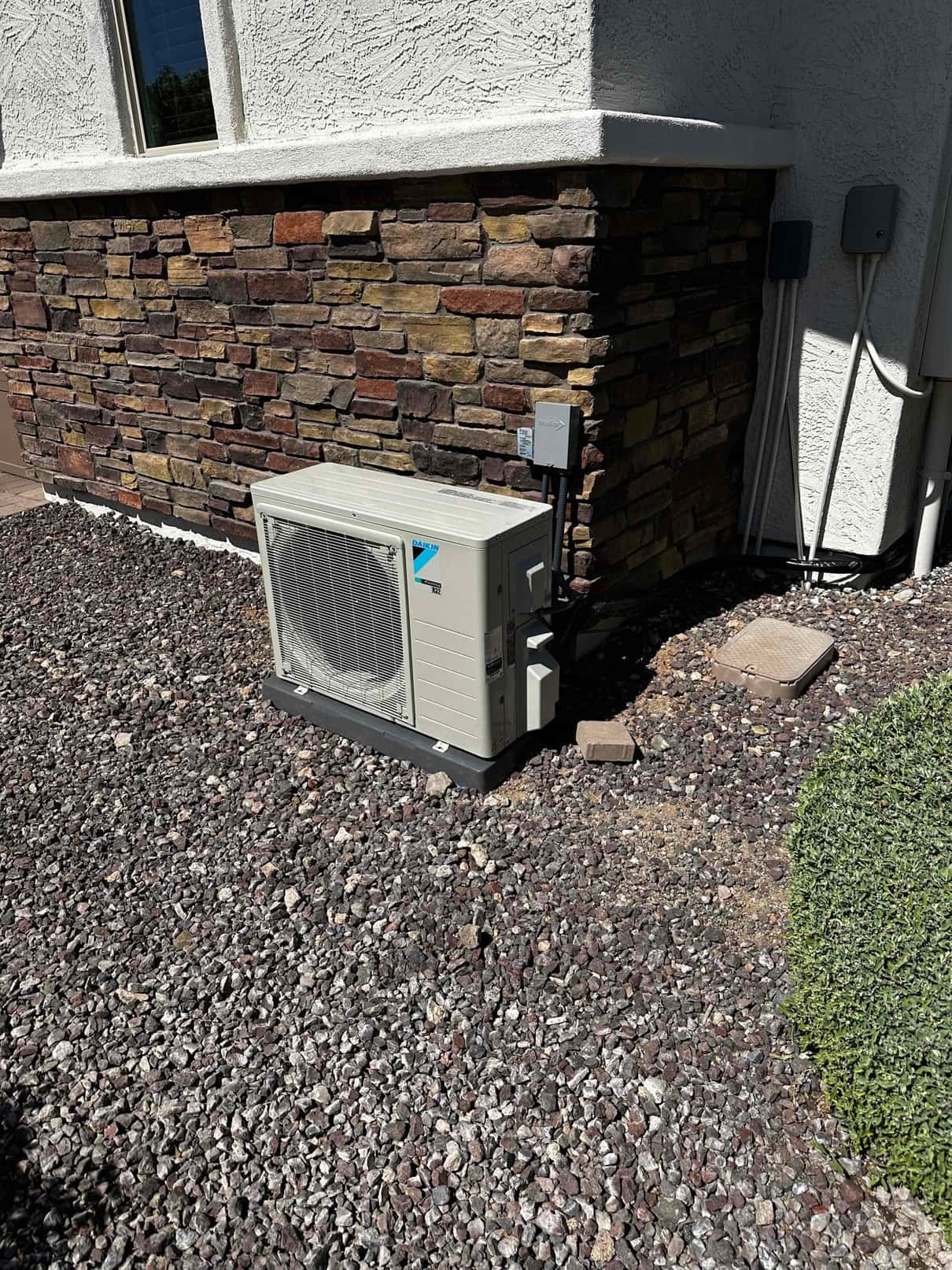 Outdoor mini-split condenser set on a composite pad with electrical disconnect and insulated line set against a stone exterior wall.