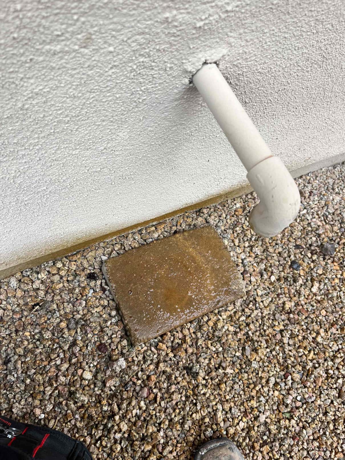 PVC condensate drain outlet terminates at exterior wall above gravel, with moisture visible on a concrete splash block.