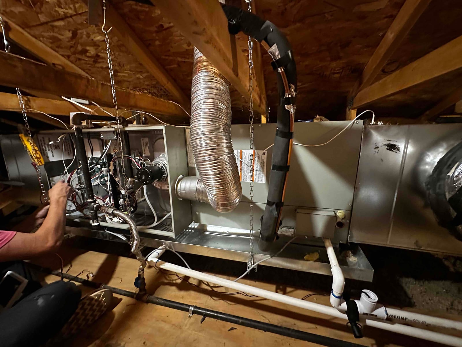 Suspended air handler installed in attic with insulated refrigerant lines, condensate drain with cleanout, and technician wiring the control compartment.