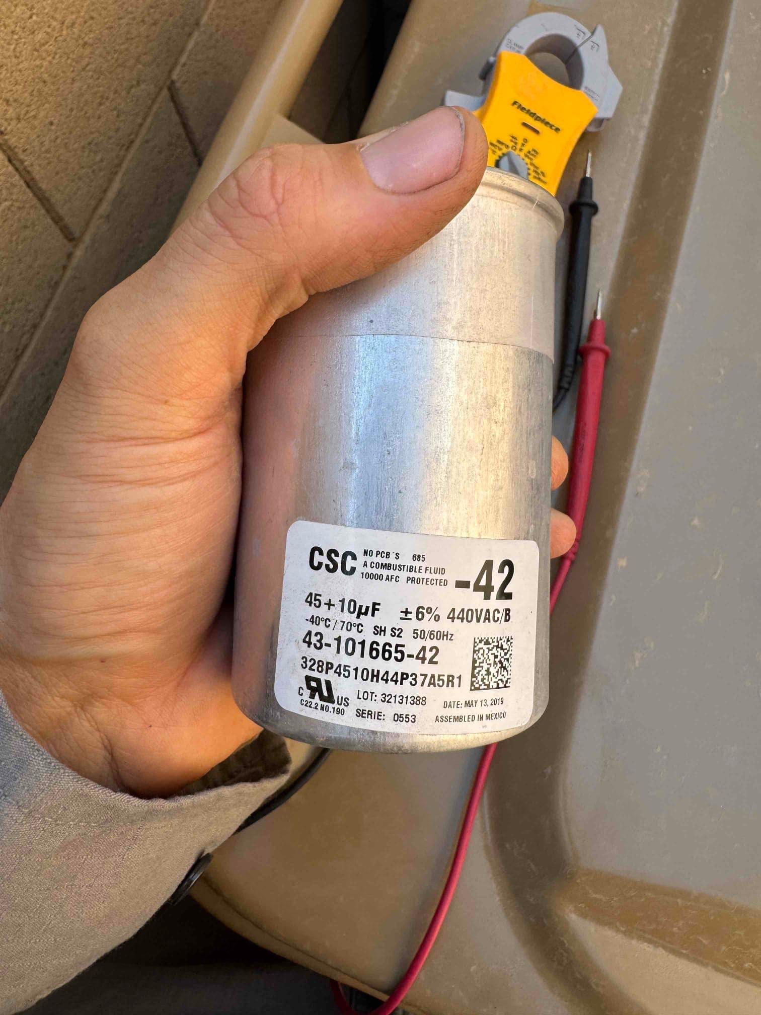 Technician holds a dual-run capacitor labeled 45+10 μF ±6% 440VAC, 40°C–70°C, removed during air conditioner repair.