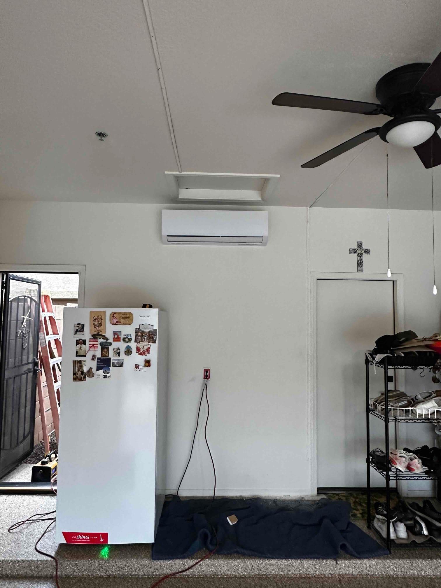 Wall-mounted mini-split indoor air handler installed in a finished garage, with nearby ladder, fan, and shelving visible.