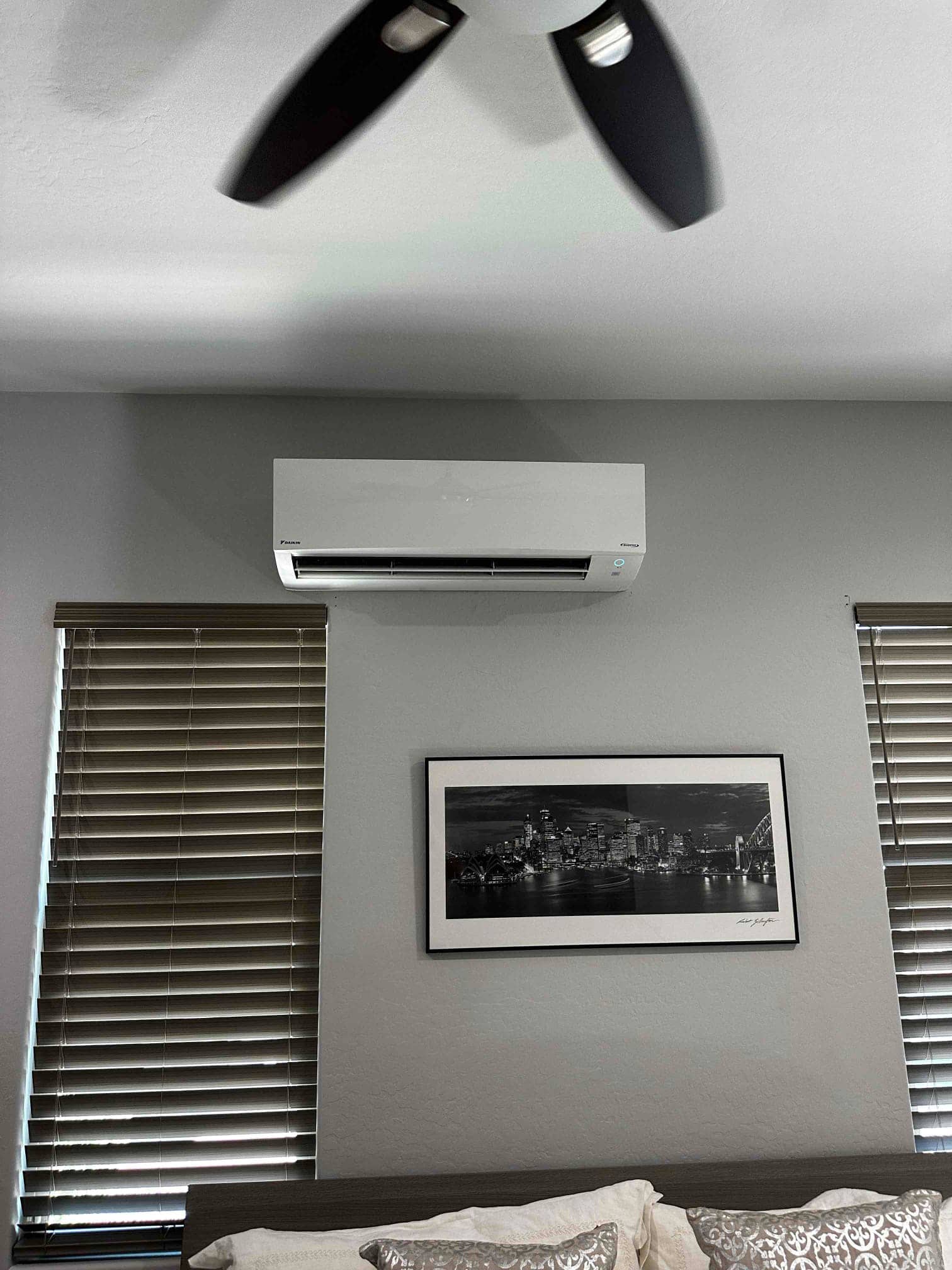 Wall-mounted mini-split indoor unit installed above a bed between two windows, operating with louvers open and indicator light on.