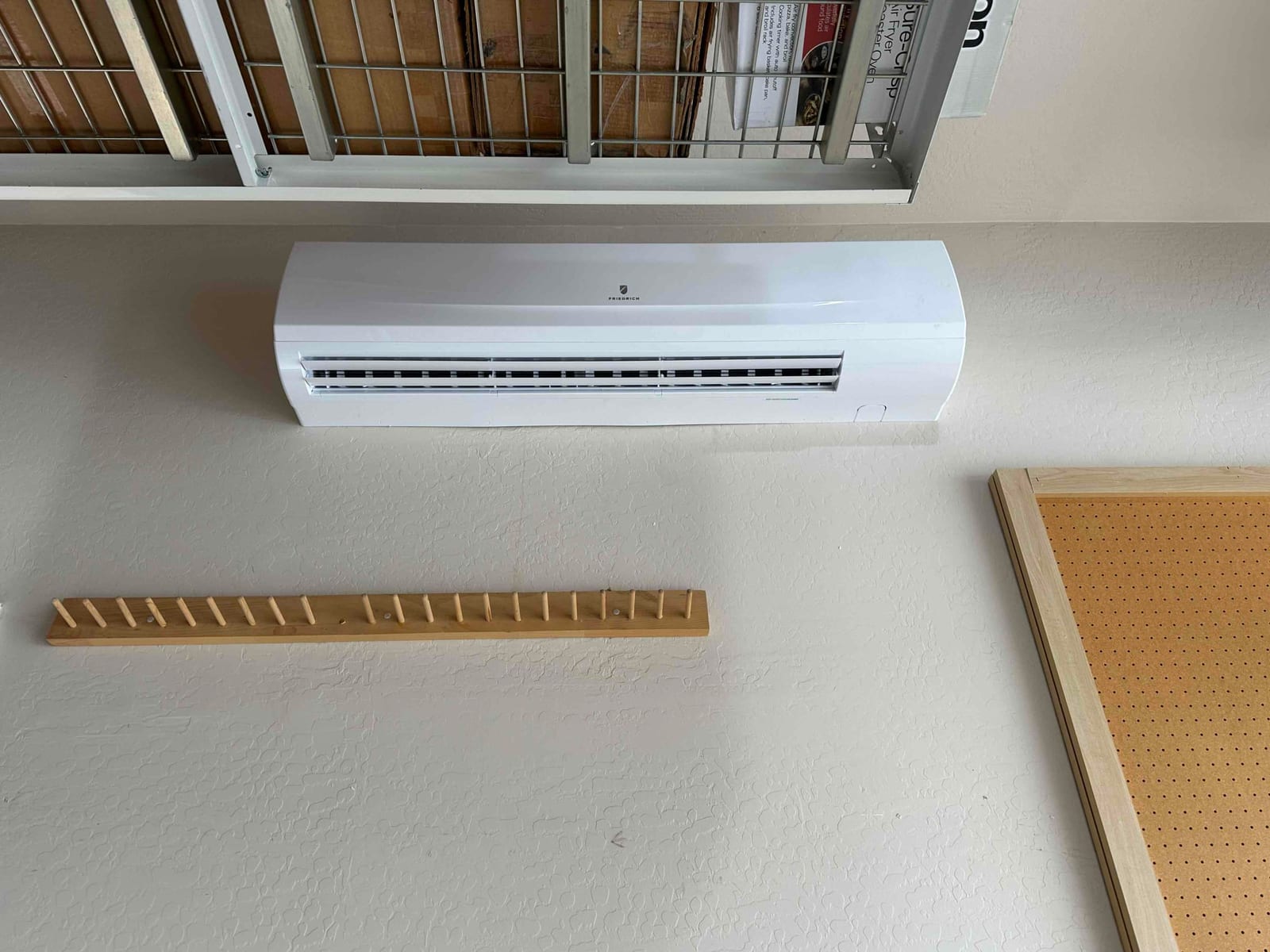 Wall-mounted mini-split indoor unit installed in a garage workspace, positioned below ceiling storage and beside a pegboard panel.