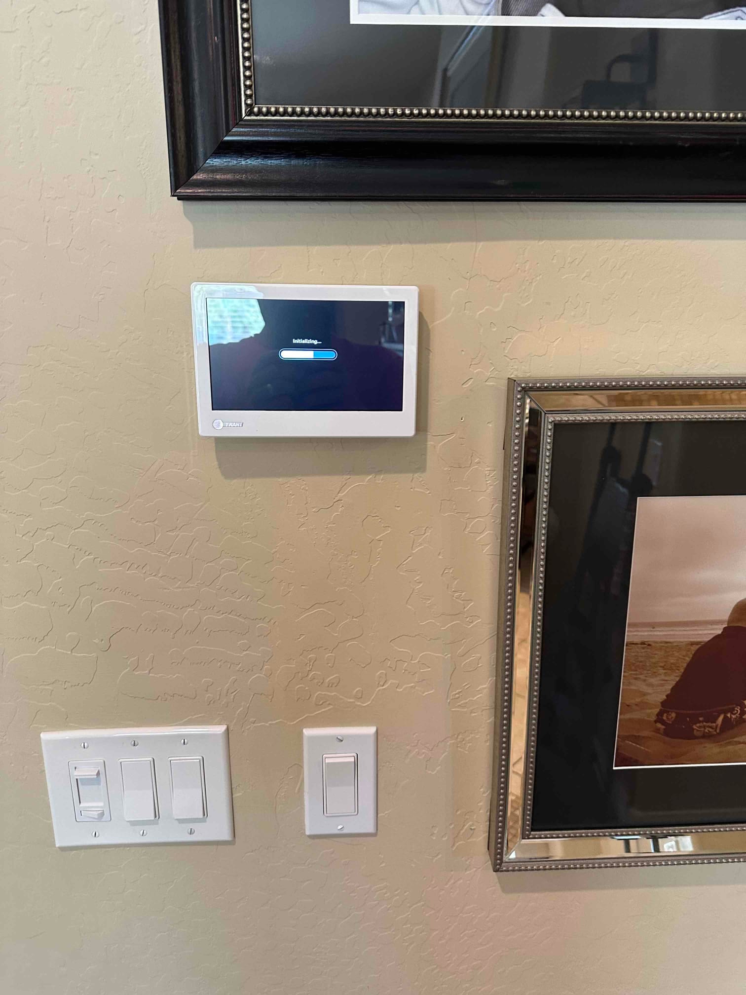 Wall-mounted touchscreen thermostat powers up and initializes after installation beside light switches and framed artwork.