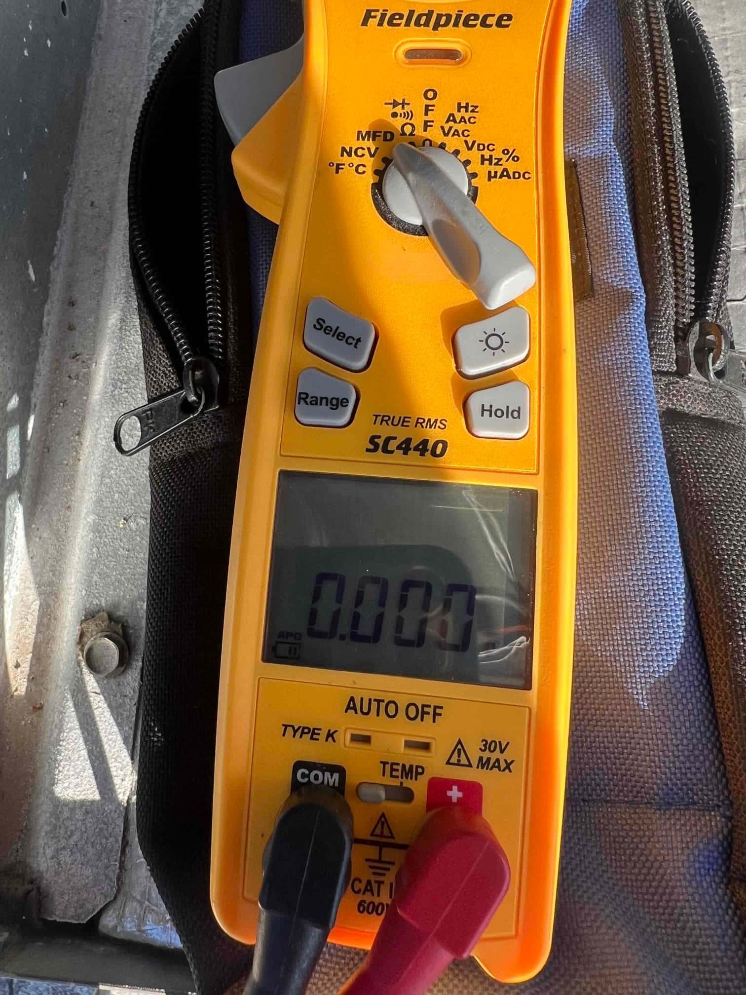 Clamp meter positioned at the outdoor unit with leads connected, display showing 0.000 and selector on AC volts mode.