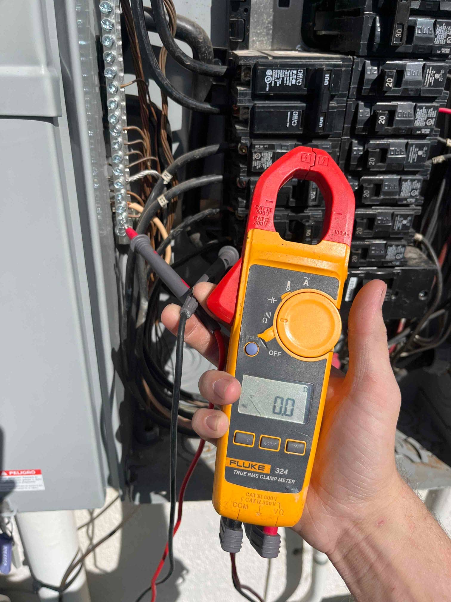 Clamp meter shows 0 A with the jaw around a conductor at the electrical panel during diagnostic testing.