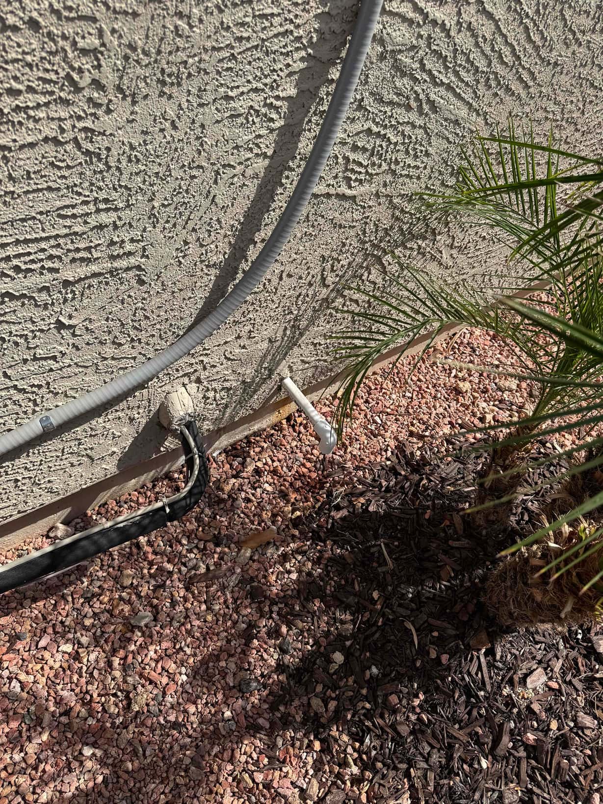 Condensate drain line termination at exterior wall above landscape rock, showing clean PVC outlet and surrounding insulated refrigerant line.