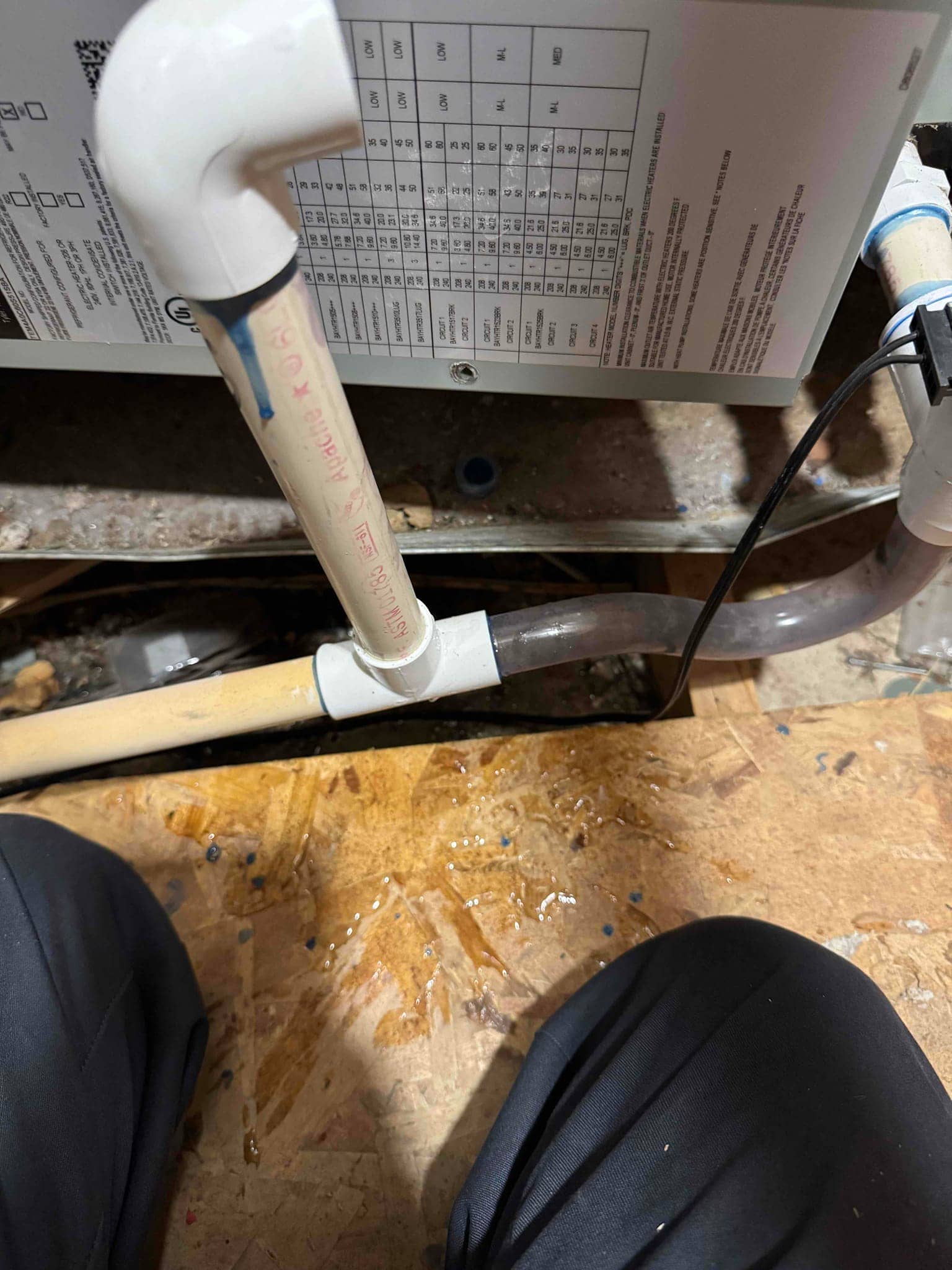 Condensate drain piping beneath an air handler shows moisture on the floor, with PVC fittings and clear trap tubing visible.