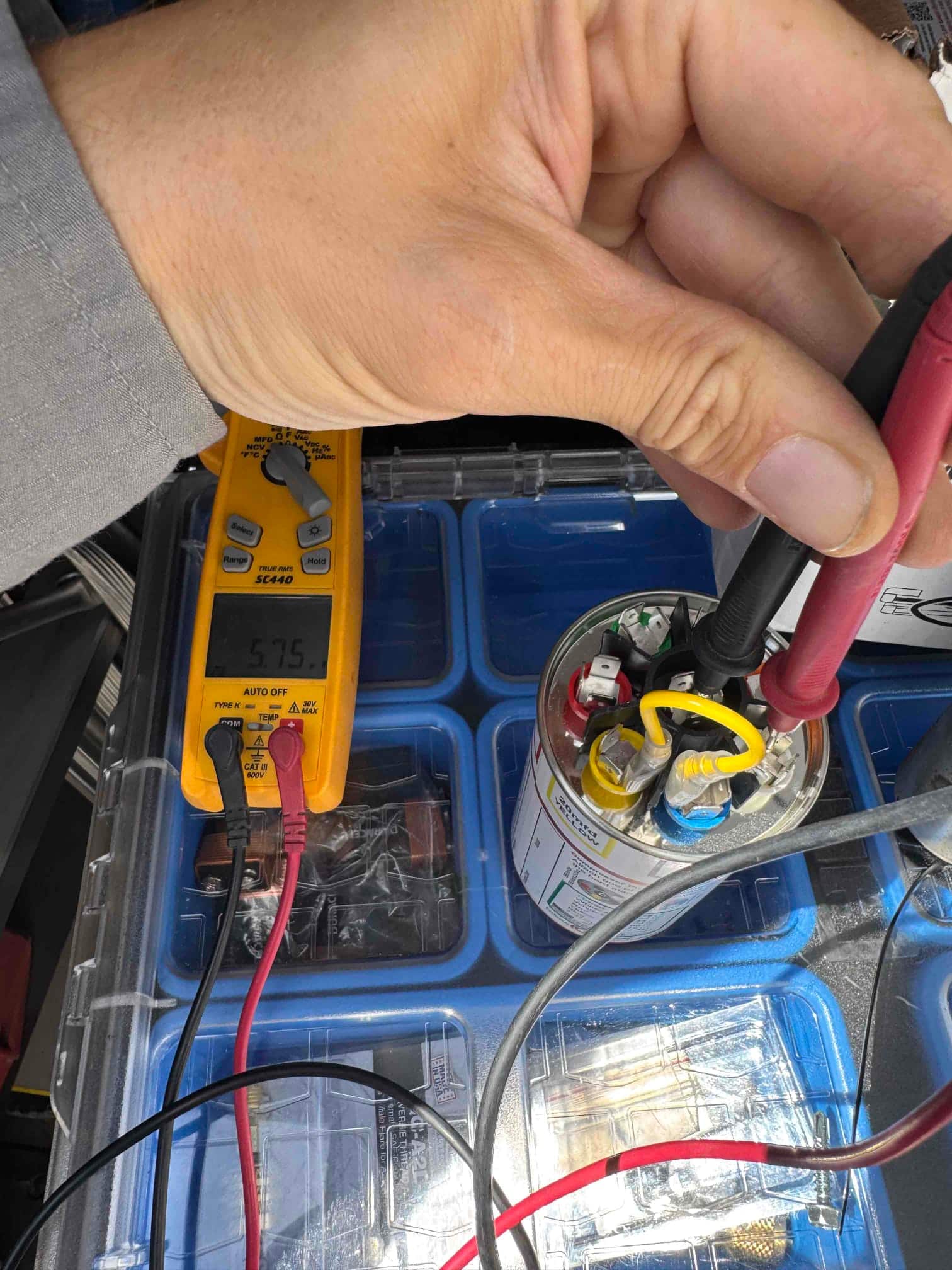 Digital multimeter measures 57.5 μF while test leads contact the terminals of a dual-run capacitor on the bench.
