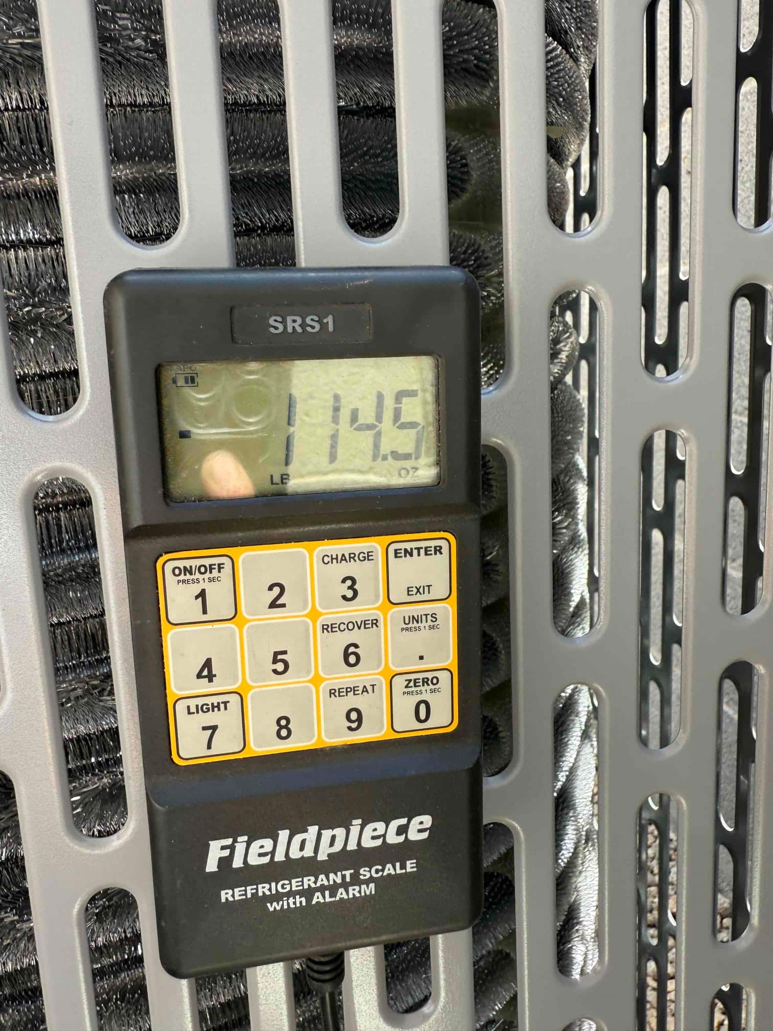 Digital refrigerant scale shows 114.5 ounces during charging at the outdoor condensing unit grille.