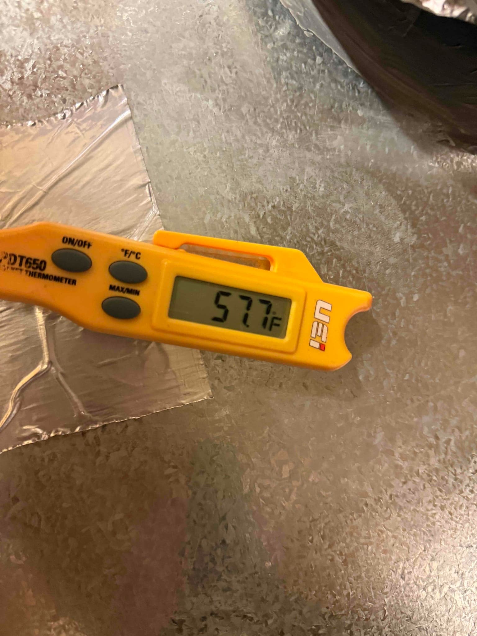 Digital thermometer shows 57.7 °F surface temperature on the metal duct or equipment panel during diagnostic measurement.