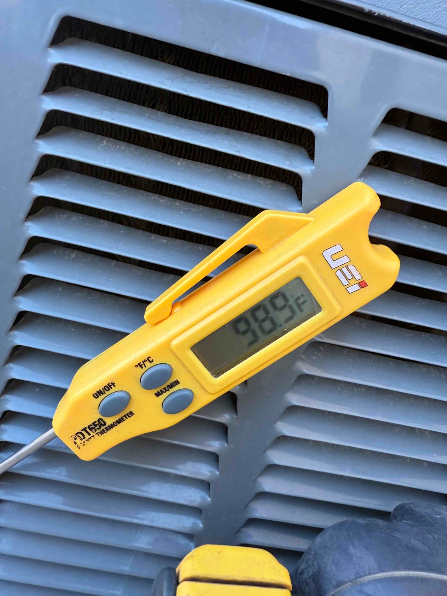 Digital thermometer shows 98.9 °F on a temperature probe placed against HVAC equipment louvers.