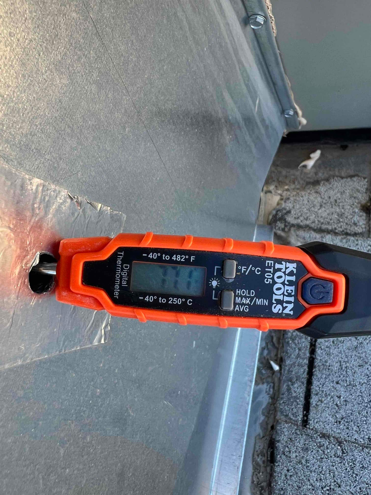Digital thermometer with surface probe attached to metal HVAC panel, showing measurement mode but the display reading is not legible.