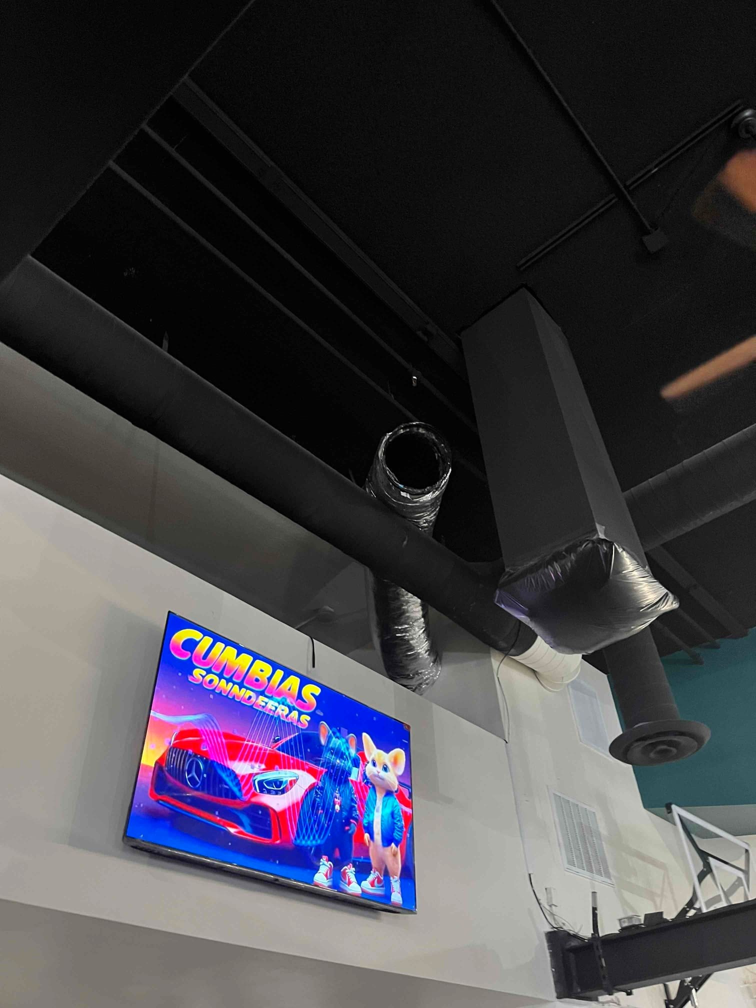 Exposed black flexible duct with open end near ceiling, showing unfinished HVAC ductwork above an interior wall-mounted television.