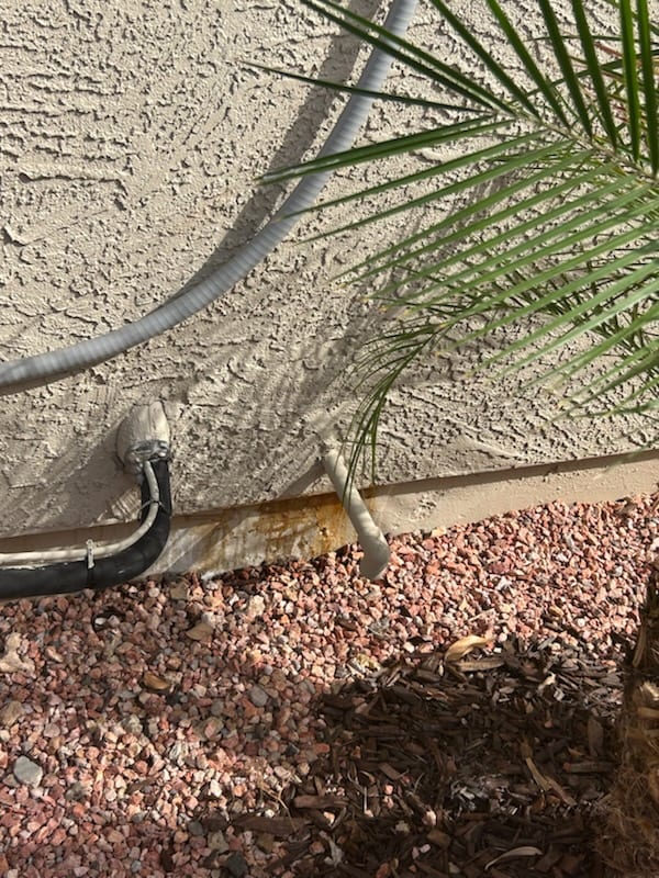 Exterior wall shows condensate drain outlet with rust staining on stucco and adjacent insulated refrigerant line above landscape rocks.