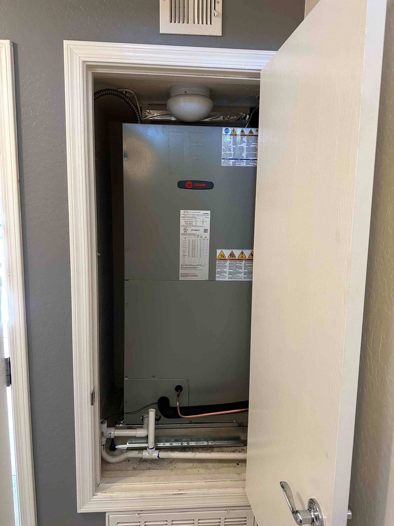 New air handler installed in a closet with insulated refrigerant lines, condensate drain piping, and return grille below the unit.