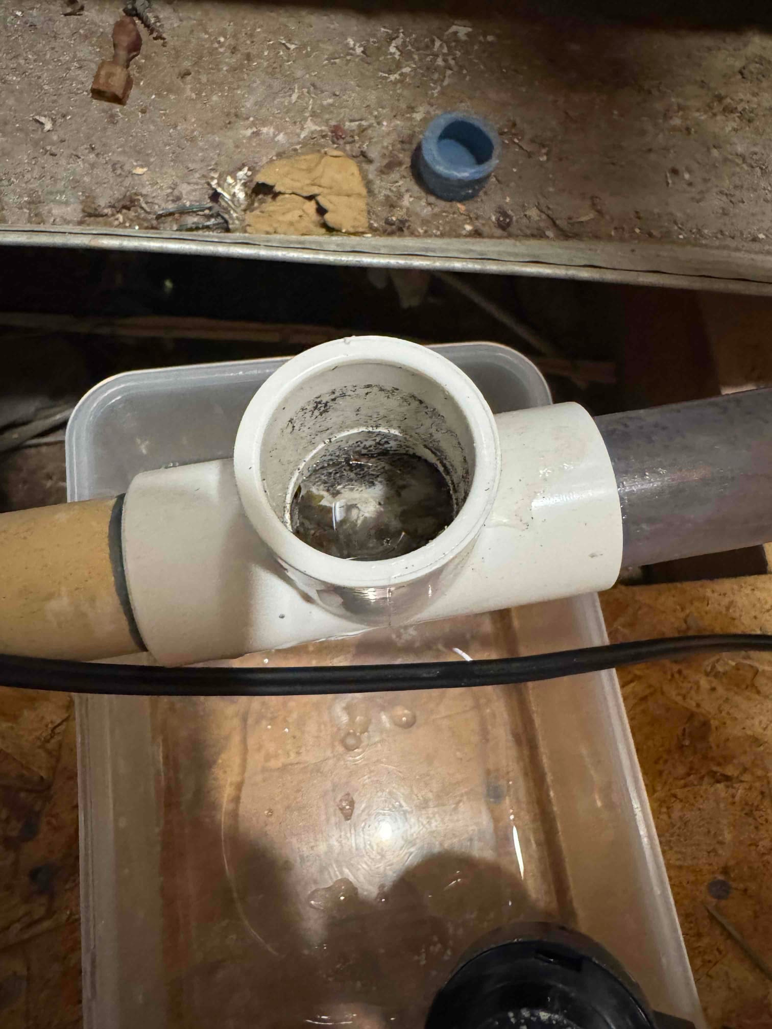 Open PVC tee on an air conditioner condensate drain line shows sludge buildup, with a catch pan below collecting water during cleaning.