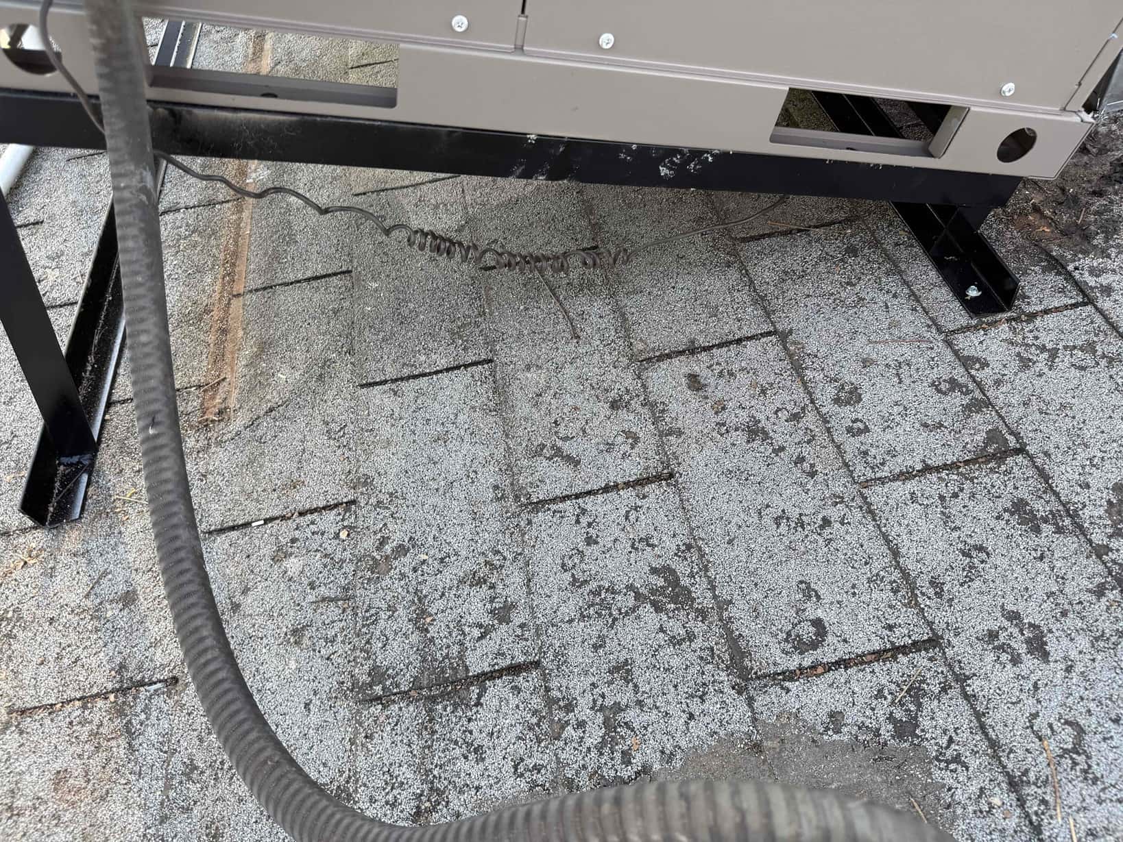 Package HVAC unit set on a raised metal stand on a shingle roof, with fasteners and conduit visible beneath.