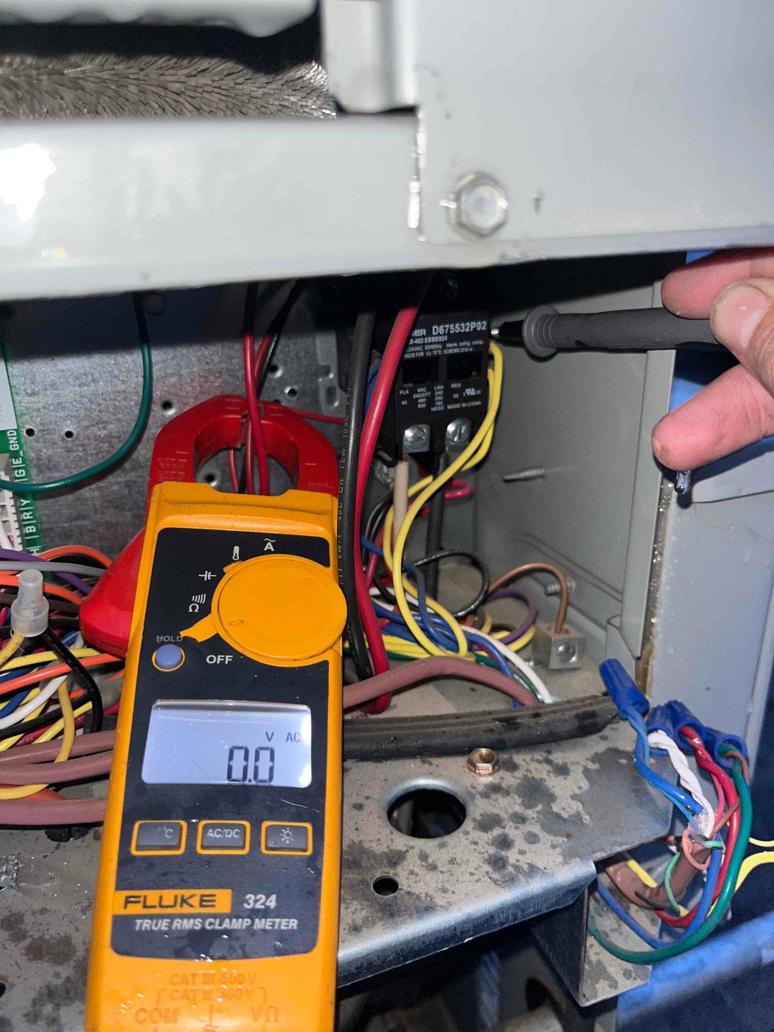 Clamp meter set to volts shows 0.0 V while probing the contactor terminals inside the condenser control compartment.