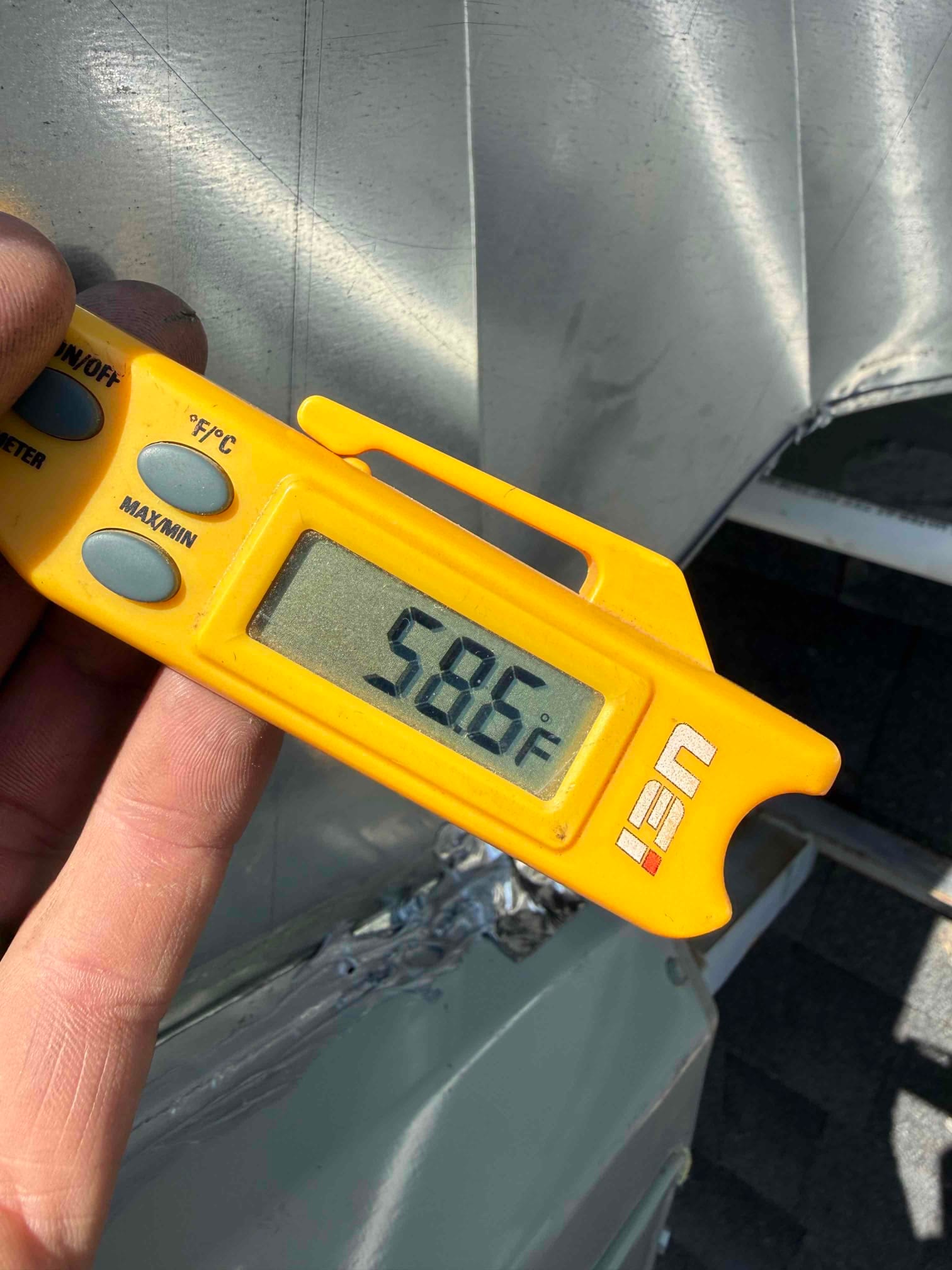 Digital thermometer shows 58.6°F while measuring air temperature at the metal duct during cooling operation.