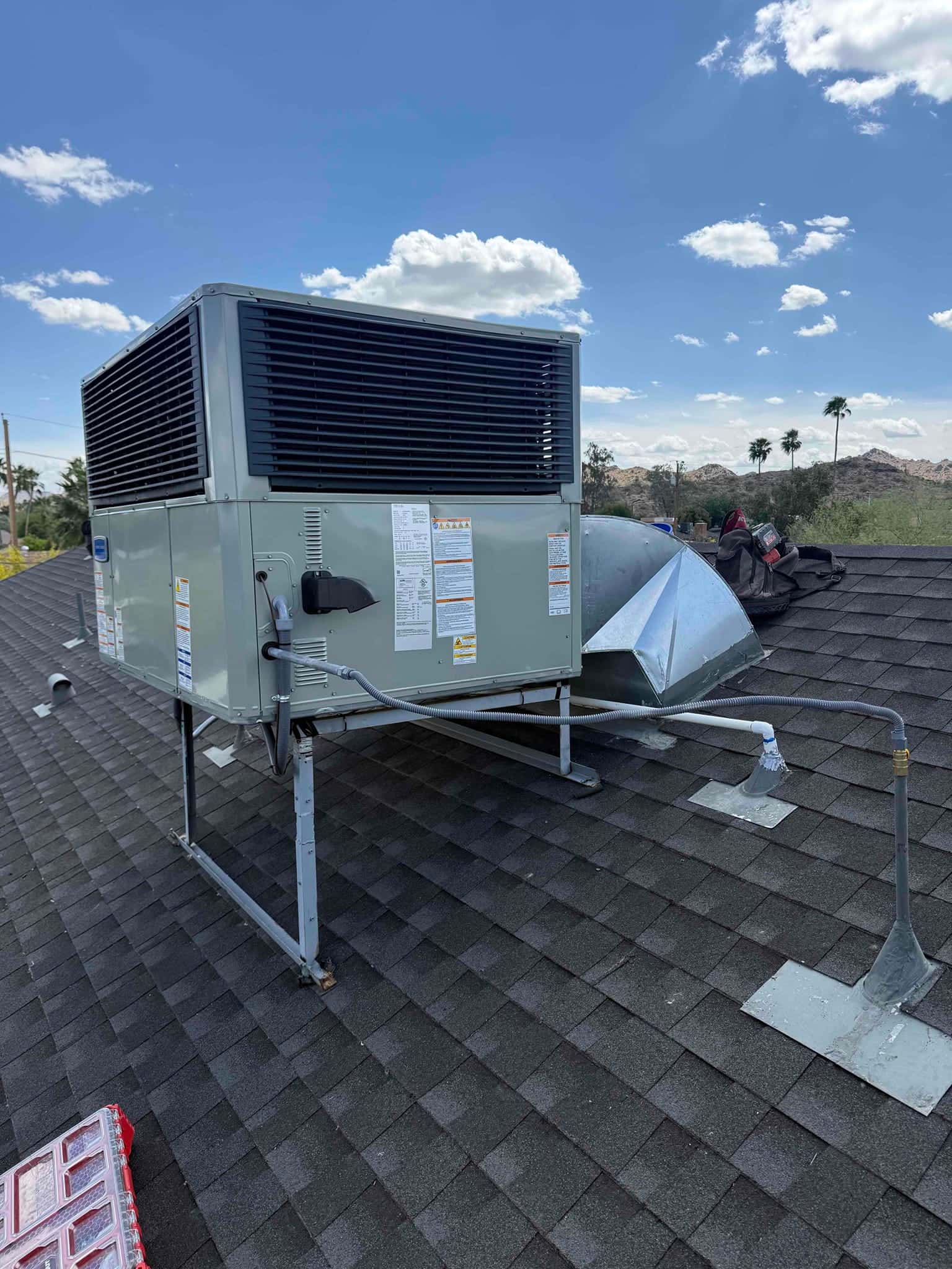 New rooftop package HVAC unit secured on a stand with connected duct transition and flexible electrical conduit on shingle roof.