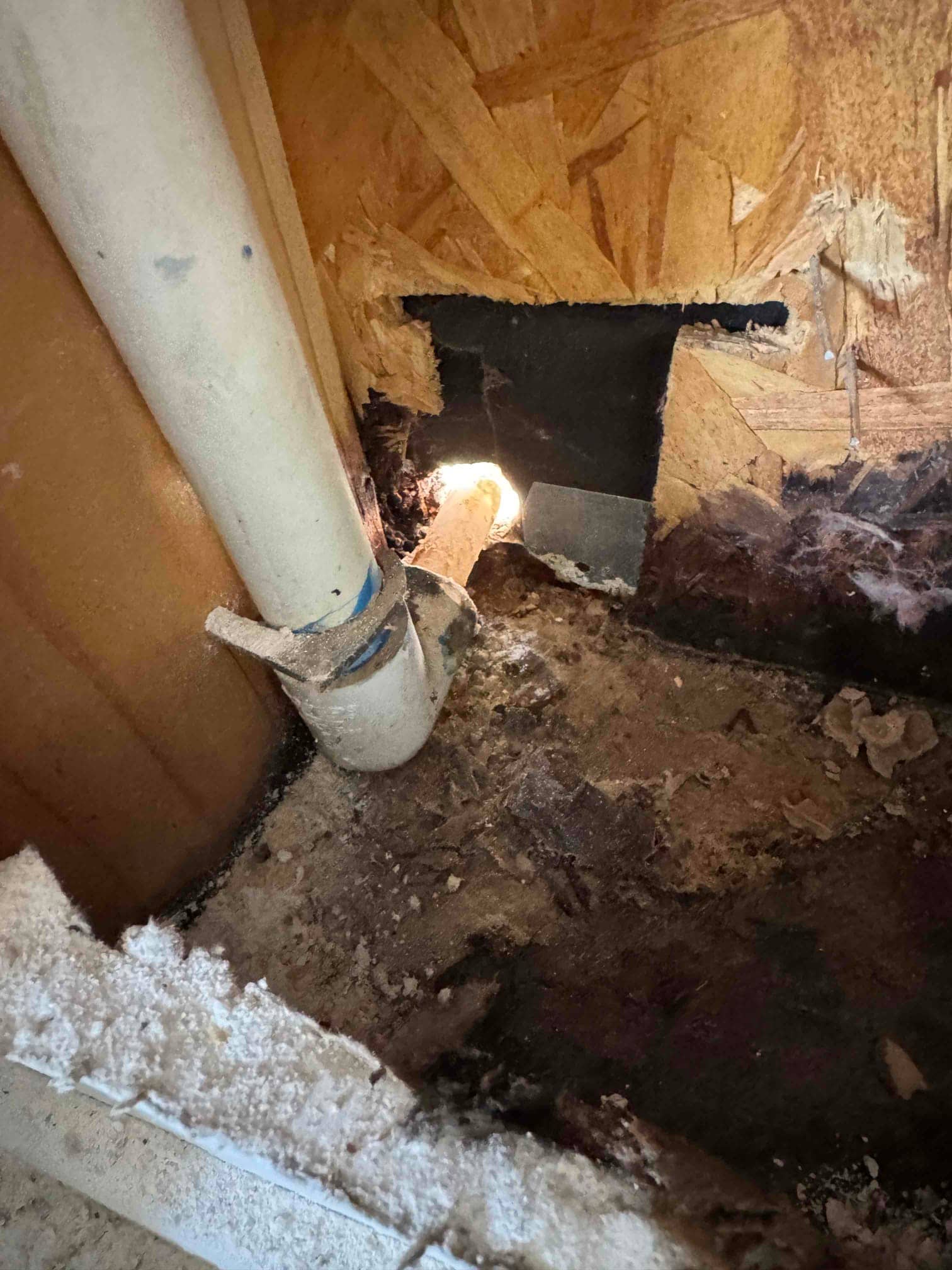 PVC condensate drain line passes through a wall cutout, showing surrounding water damage, debris, and deteriorated subfloor near the drain.