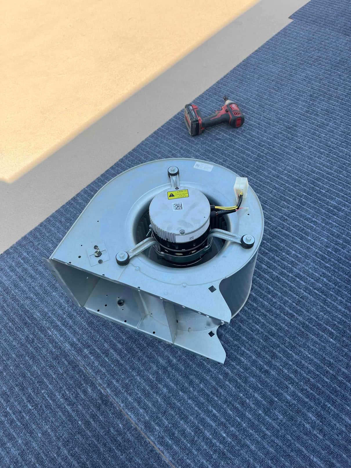 Removed indoor blower assembly with replacement motor installed, showing wiring harness and housing on a work mat beside a drill.