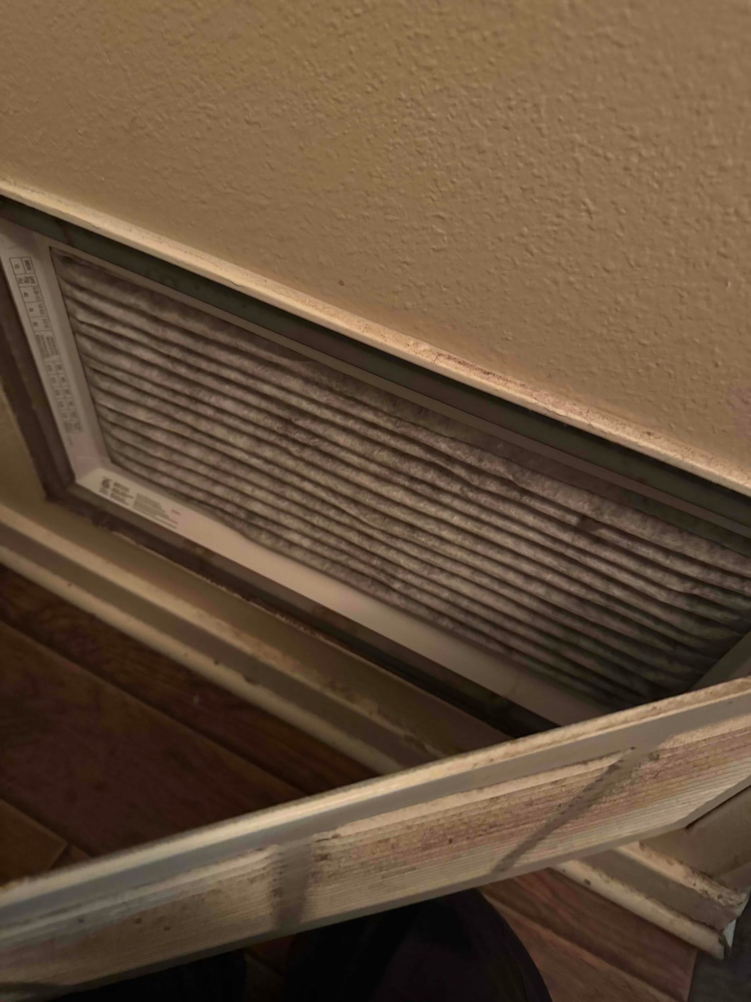 Return air filter behind a grille shows heavy dust accumulation on the pleats inside a wall-mounted intake.