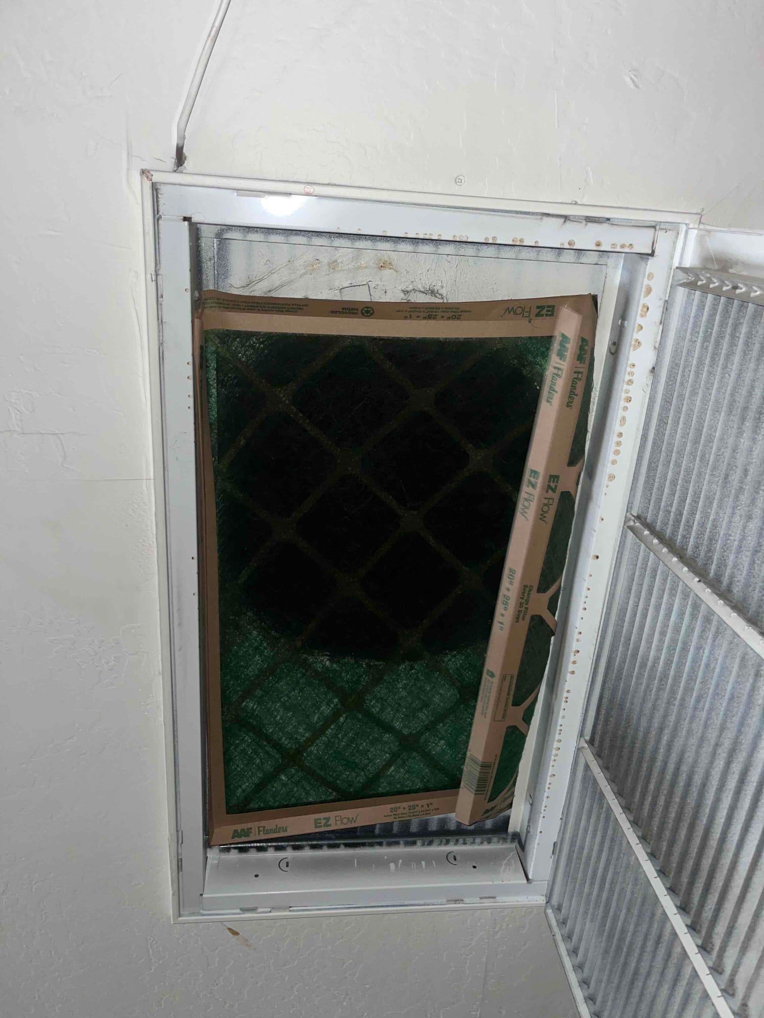 Return air grille open with a green pleated air filter partially inserted, showing frame edge and surrounding grille frame.