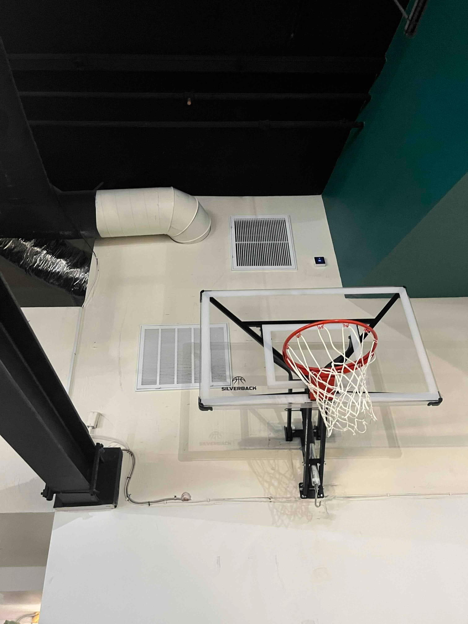 Return air grilles and ductwork on an indoor wall near a mounted basketball hoop in a gym space.