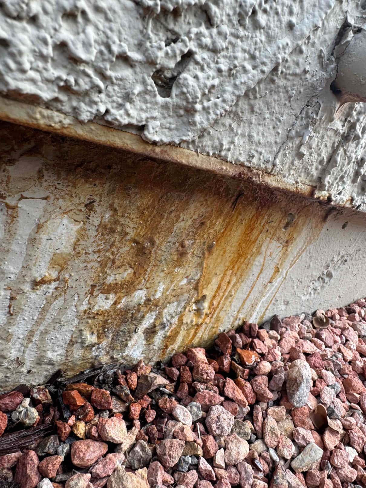 Rust-stained exterior wall and foundation with mineral deposits beneath a condensate drain outlet above decorative gravel.