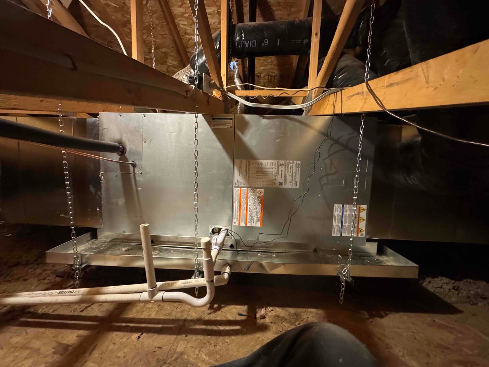 Suspended air handler installed in attic with insulated refrigerant line, condensate drain piping, and overflow pan supported by chains.