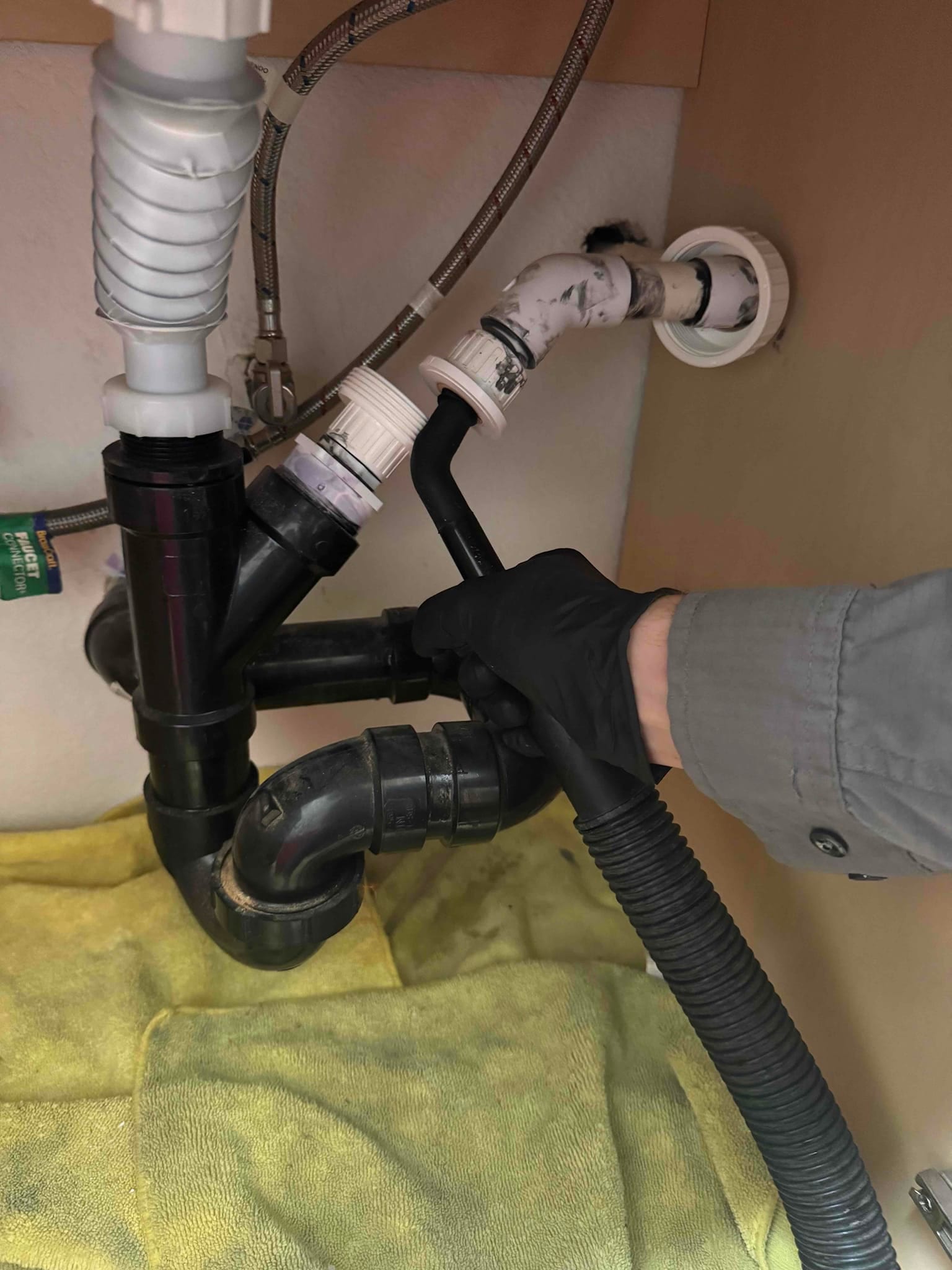 Technician’s gloved hand clears a flexible condensate drain line connection under a sink, showing PVC traps and fittings.