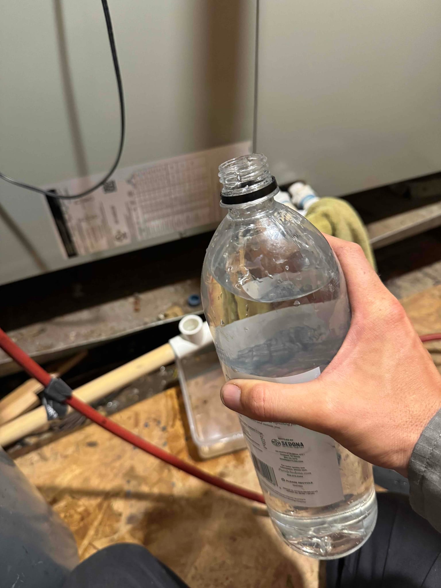 Technician’s hand holds a water bottle near an exposed condensate drain line and trap at the air handler.