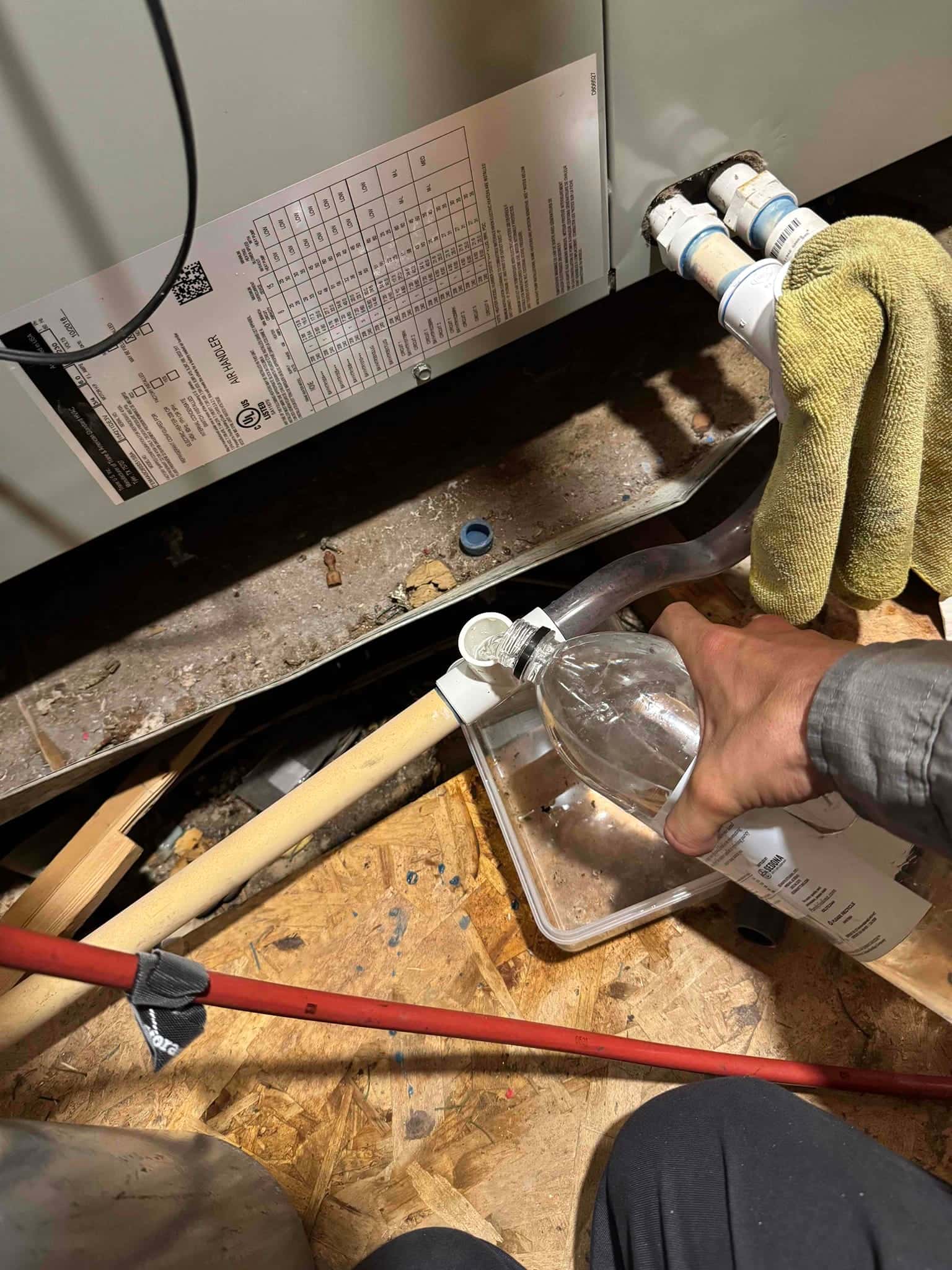 Technician pours cleaning solution into an air handler’s condensate drain line, with PVC fittings and drain pan visible.