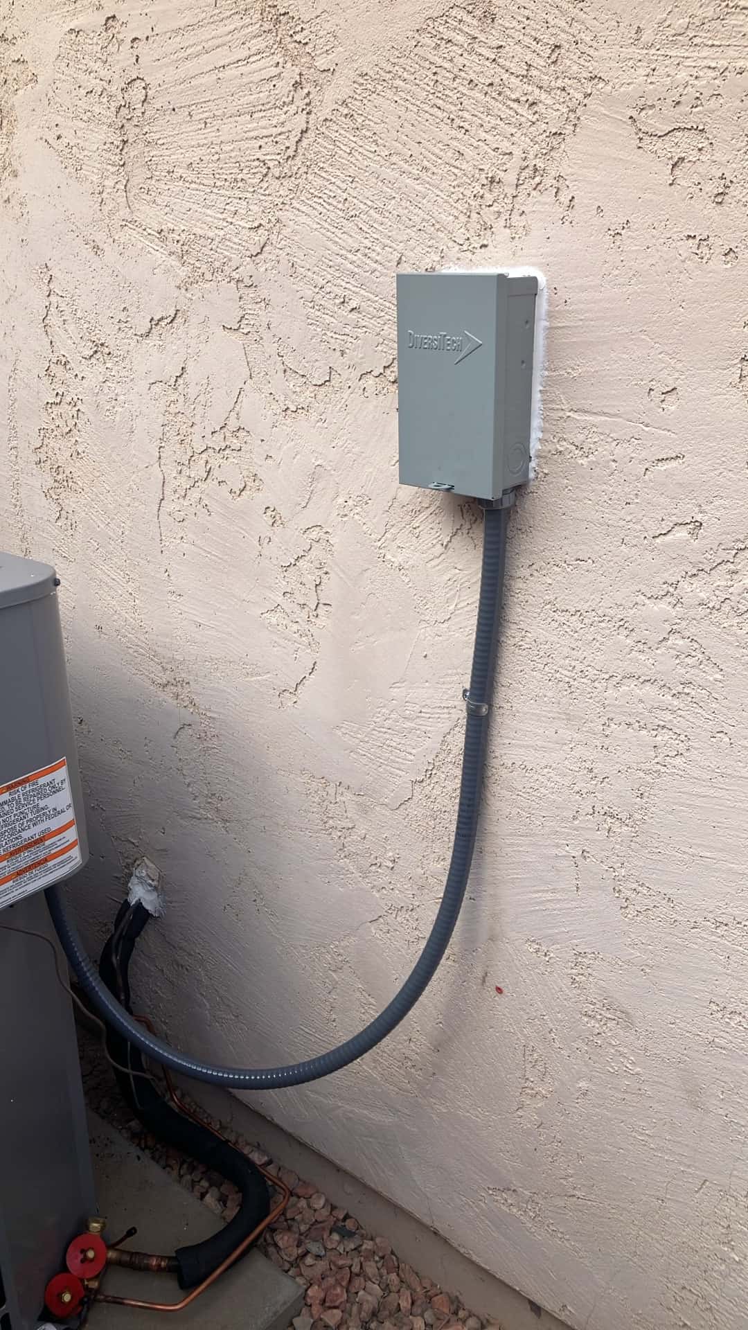 Weatherproof HVAC disconnect mounted on stucco wall with sealed penetration and flexible conduit feeding the new outdoor unit.